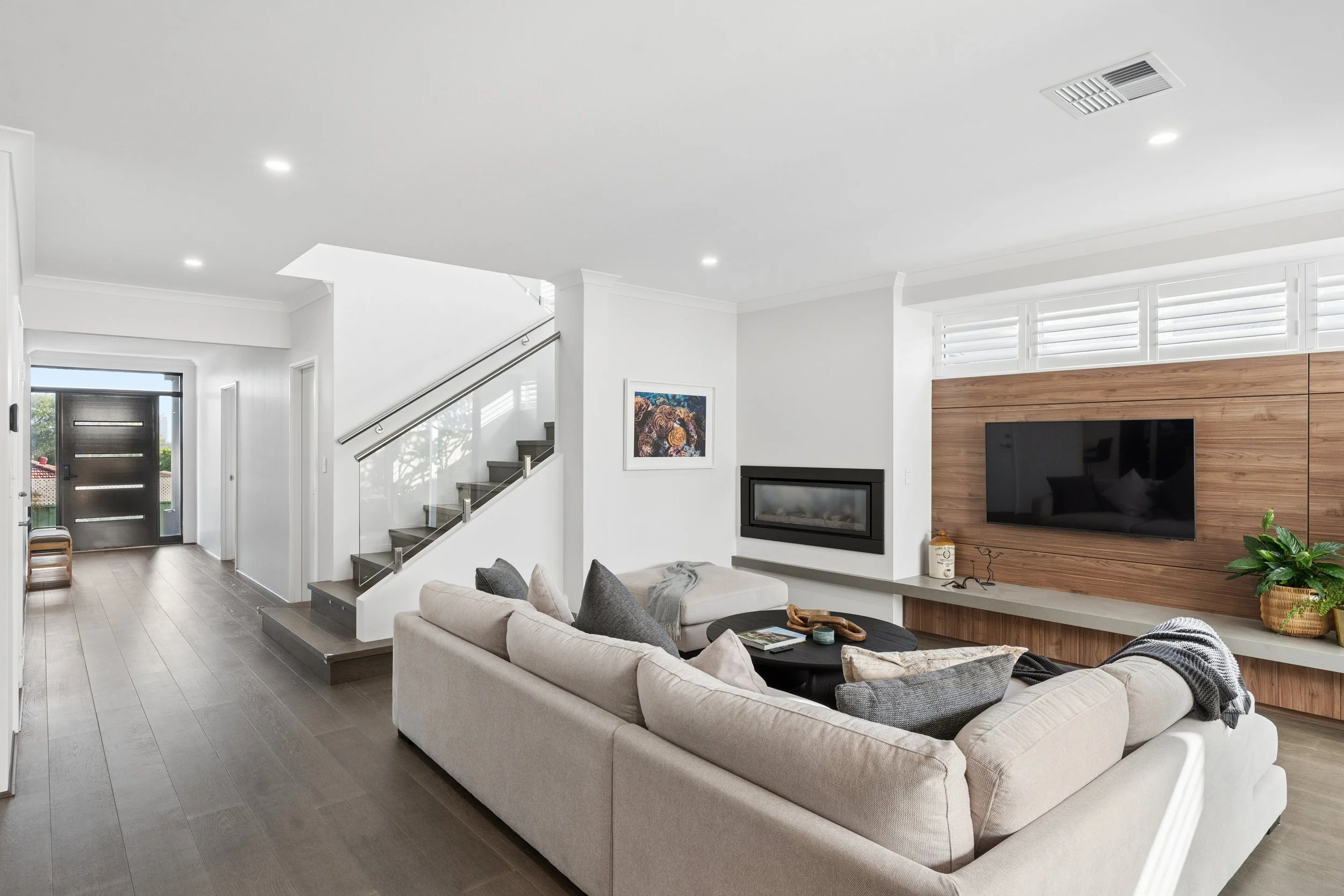 Modern living room with white walls, a light-colored sofa, a fireplace, and a flat-screen TV mounted on a wooden accent wall. Large windows with white plantation shutters allow natural light.