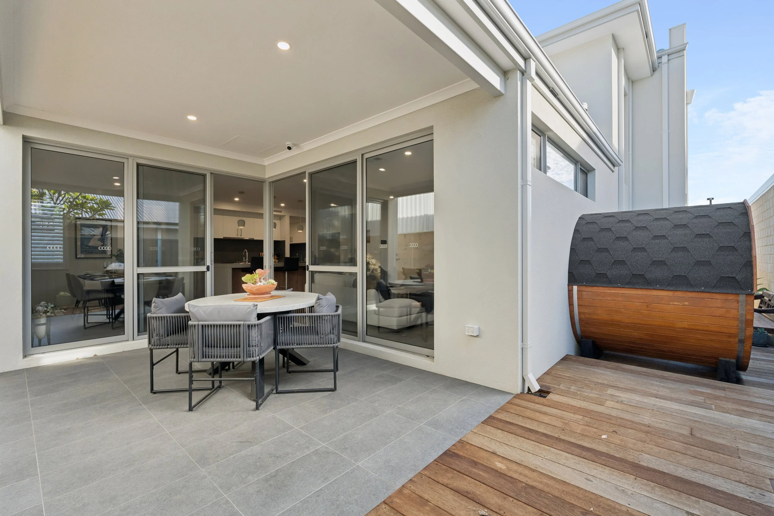 Modern house exterior with glass sliding doors leading to a patio, outdoor dining table with chairs, and a wooden deck area with a small storage shed.