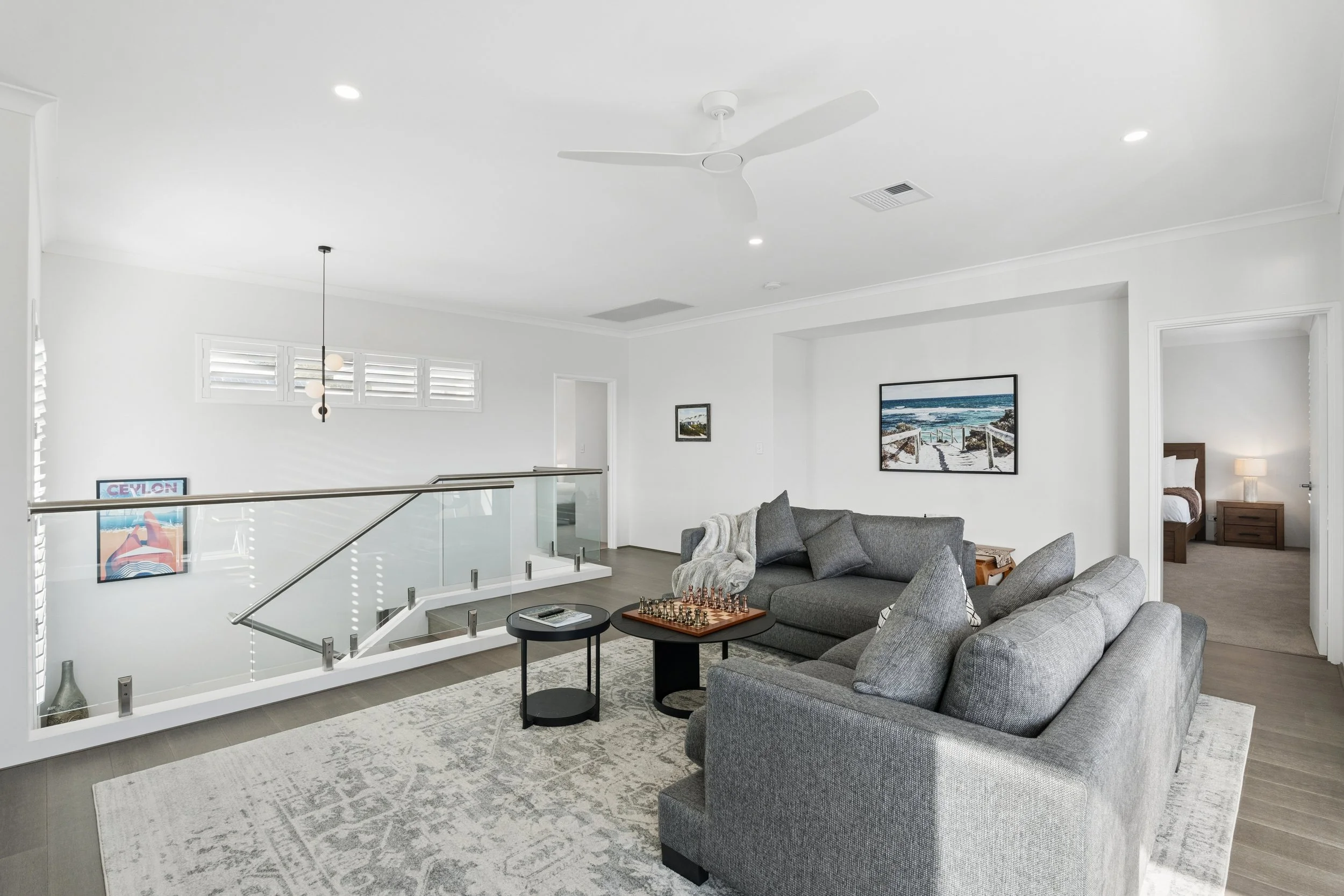 Modern living room with gray sofas, a round black coffee table with a chess set, framed artwork on white walls, and large windows with shutters.
