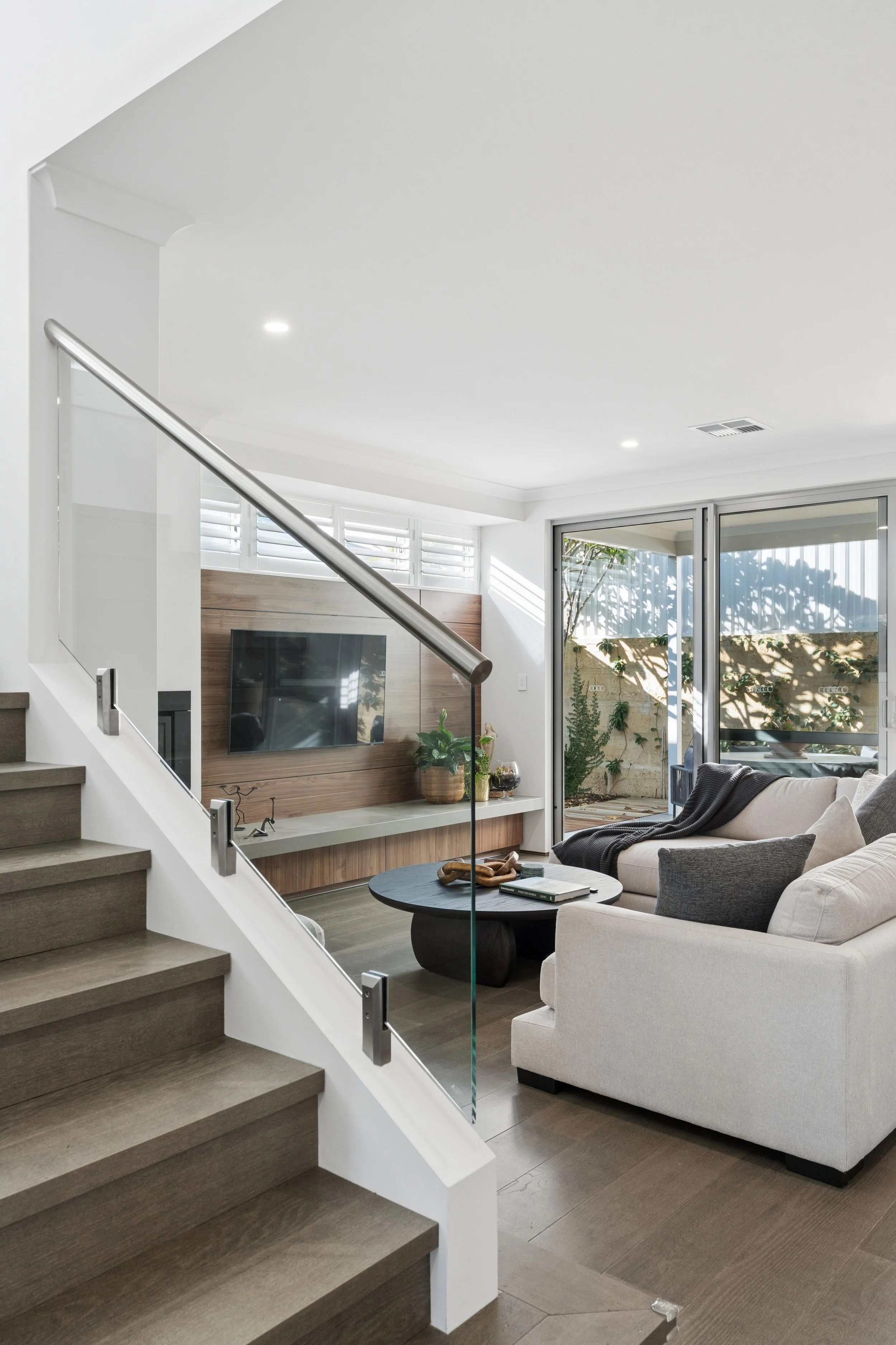 Modern living room with a staircase on the left side, a large window with white plantation shutters and sliding glass door on the right, a light-colored sofa, a round black coffee table, a wall-mounted television, wood paneling, and indoor plants.