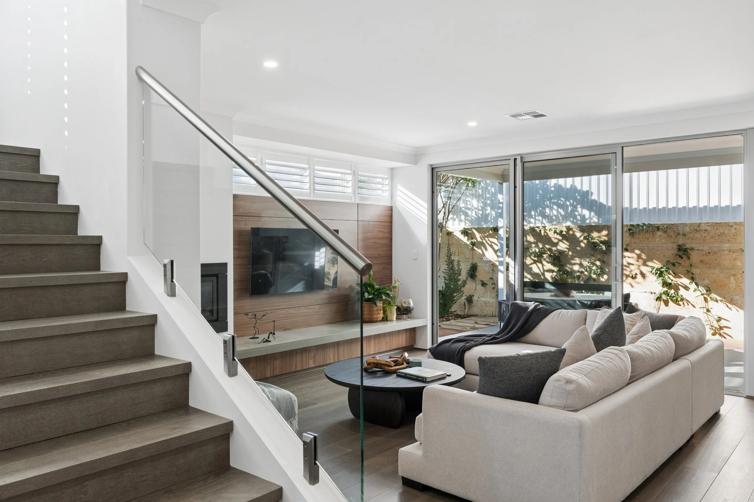 Modern living room with beige sofa, black coffee table, and sliding glass door leading to outdoor patio with plants.