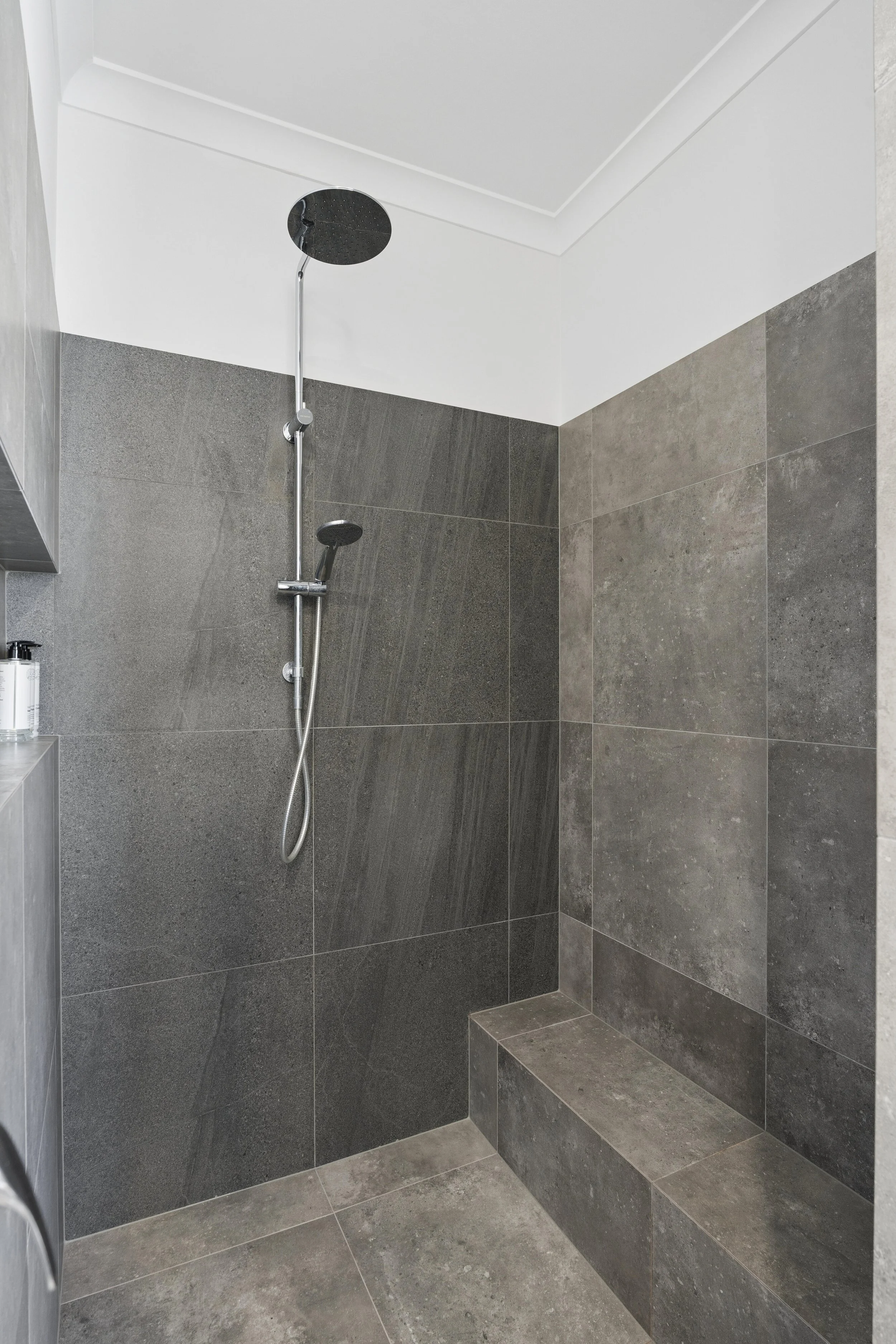 Interior of a modern shower with large grey tiles, a built-in bench, and a ceiling-mounted rainfall showerhead.