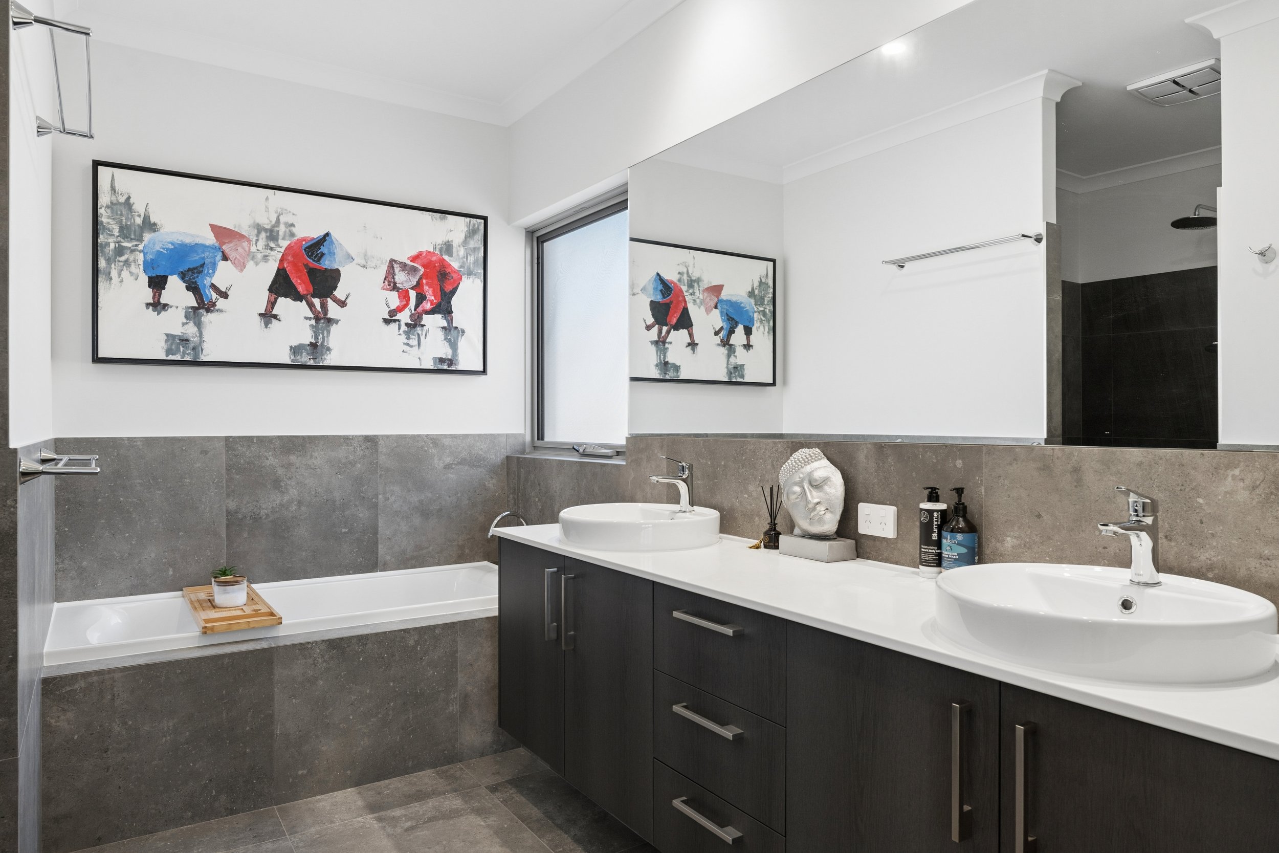 Modern bathroom with double sinks, dark cabinet, grey tiled walls, large wall mirror, decorative head sculpture, and artwork of people working in a field.