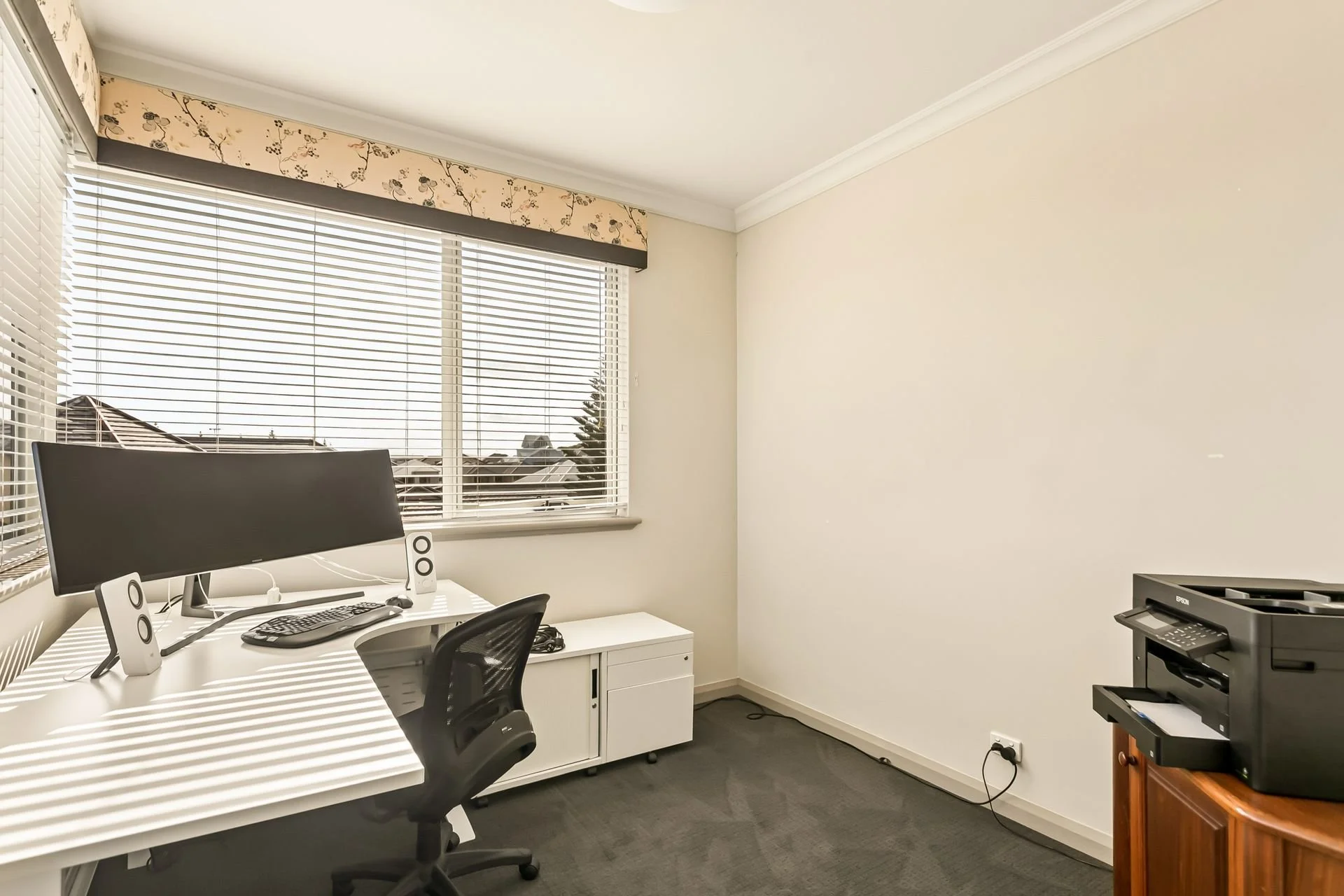 An office with a window having blinds and floral valance, a white desk with a computer monitor, keyboard, mouse, and speakers, black office chair, a small white cabinet with a printer, and a wooden table with a black printer.