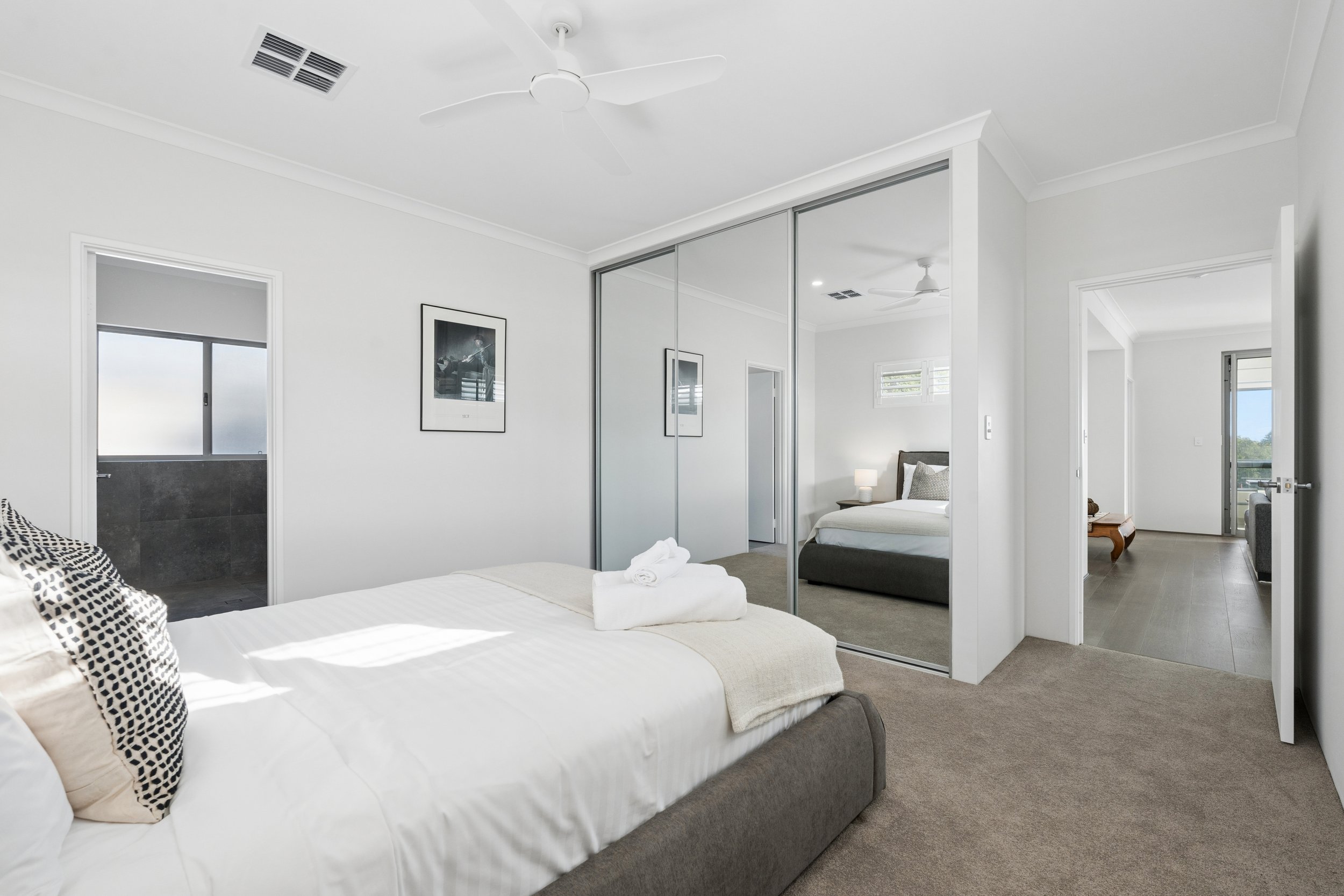 A bedroom with a bed, mirrored closet doors, and a doorway leading to a bathroom and another room, all decorated in neutral tones.