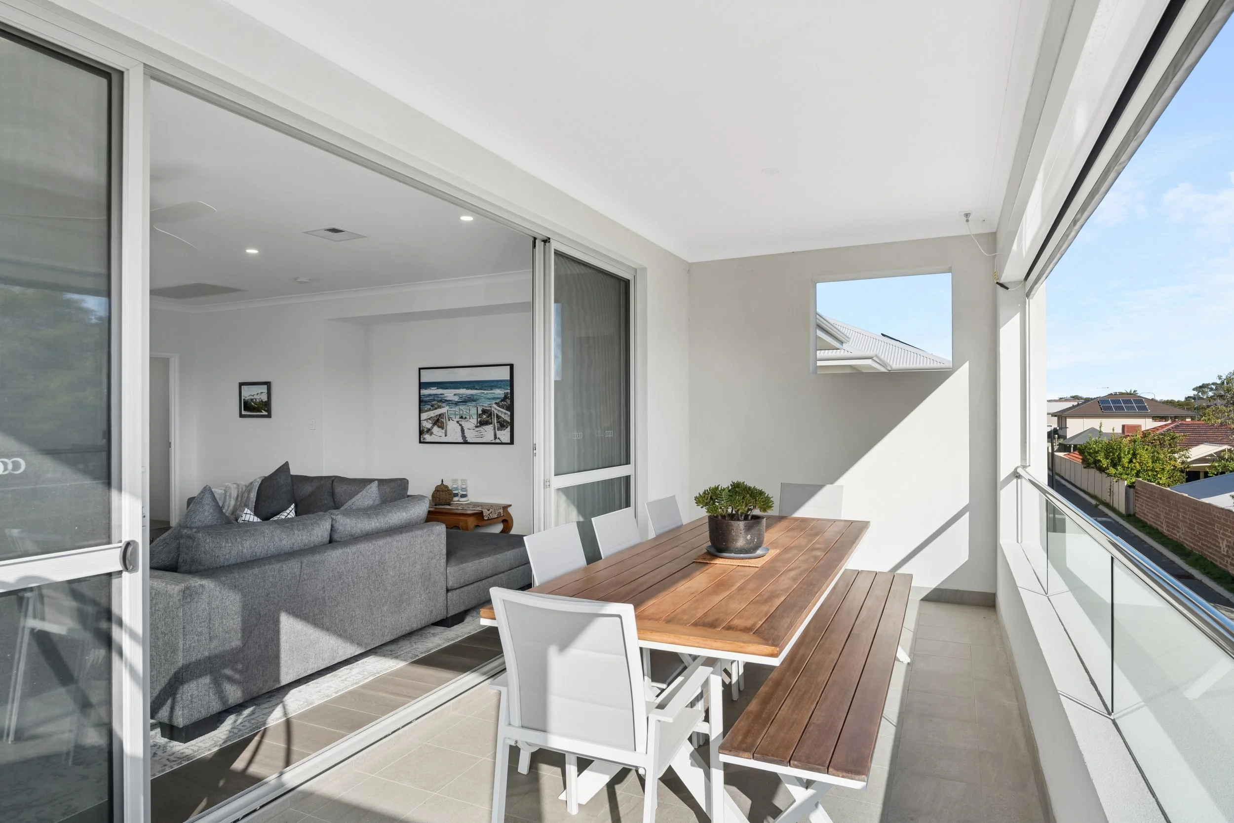 A spacious balcony with a wooden dining table and white chairs, overlooking a suburban neighborhood with houses and trees, connected to an indoor living area with a gray sofa and wall art.