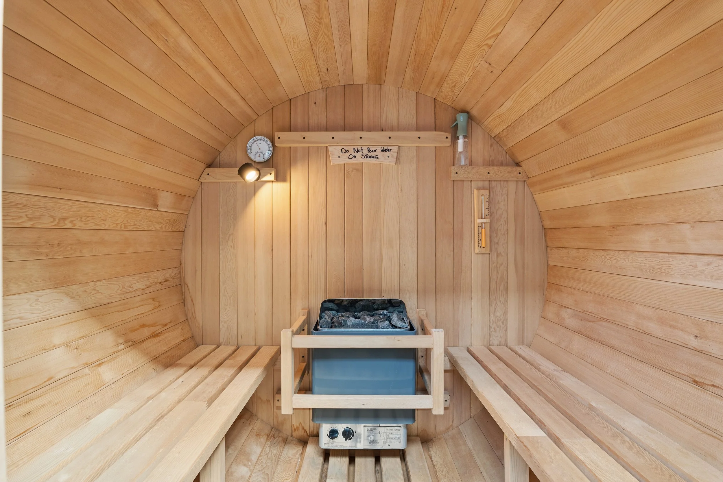 Interior of a wooden sauna with benches on either side, a heater with stones in the center, and a thermometer and a sign that reads 'Do Not Pour Water On Stones' on the wall.