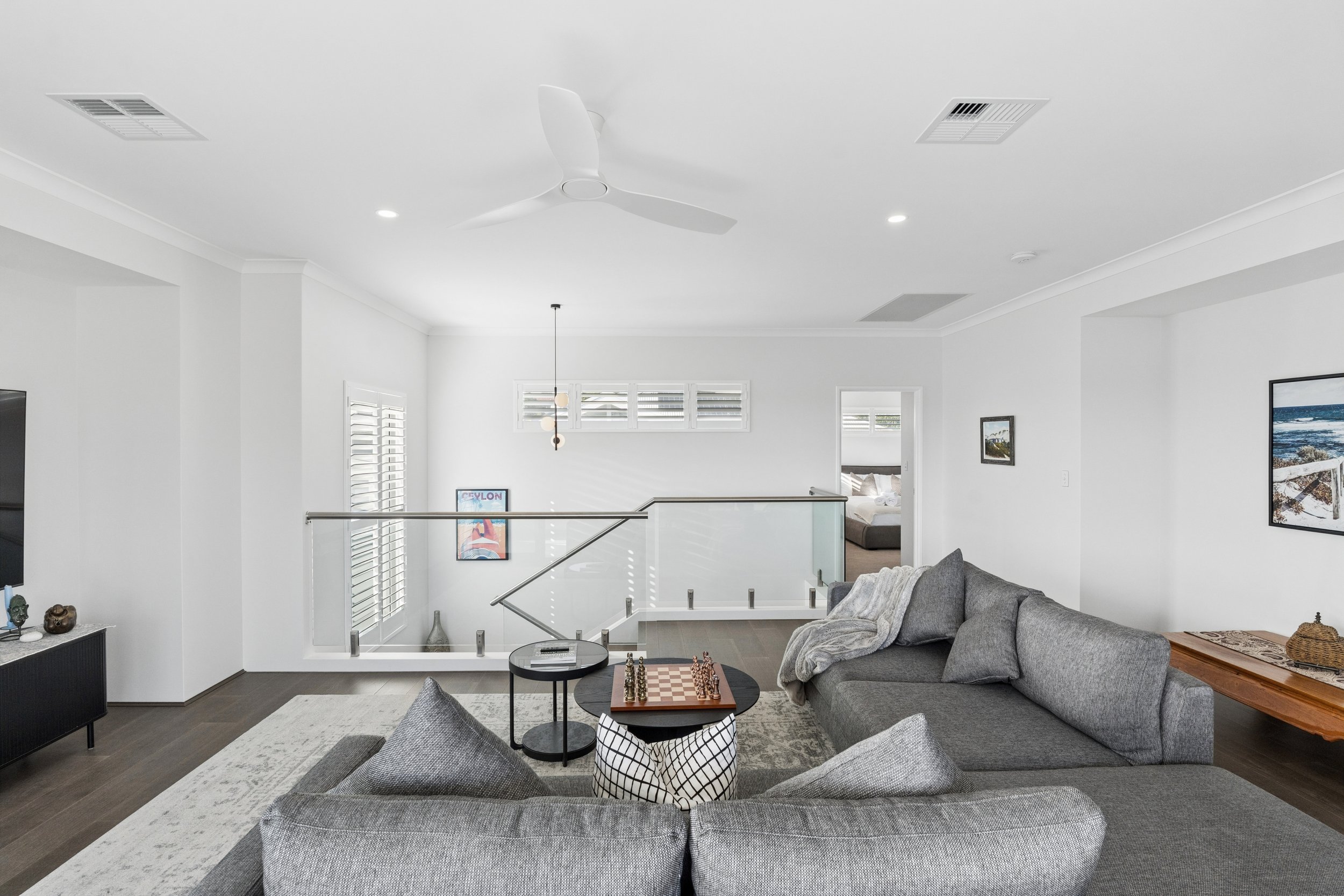 Living room with a grey sectional sofa, a round black coffee table with chess and checkers, and a wooden side table with a basket. White walls decorated with framed artwork, large windows with shutters, and a staircase railing. Ceiling fan and recess
