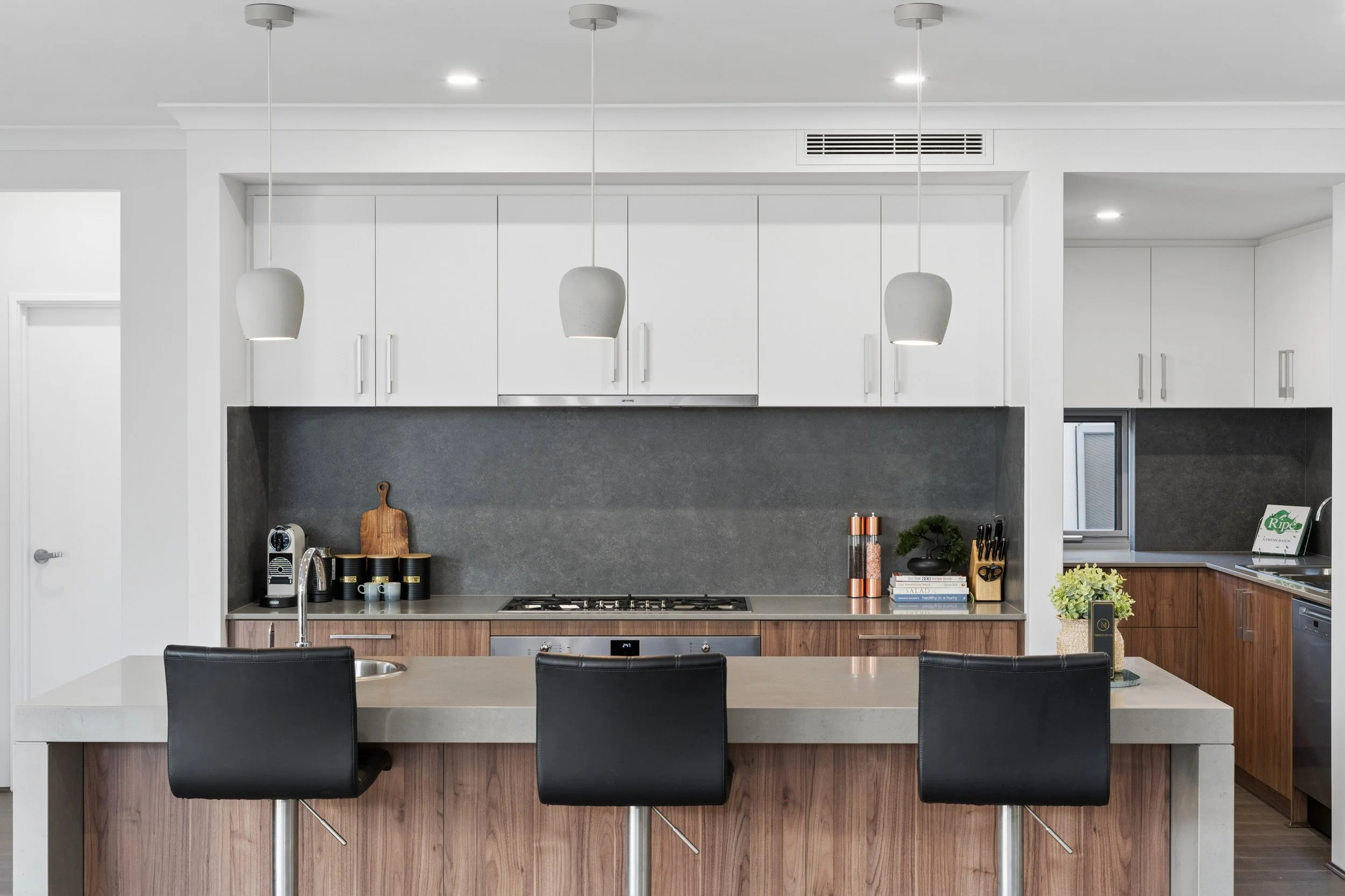 Modern kitchen with white upper cabinets, gray backsplash, and wooden lower cabinets, featuring a kitchen island with black chairs and pendant lights.