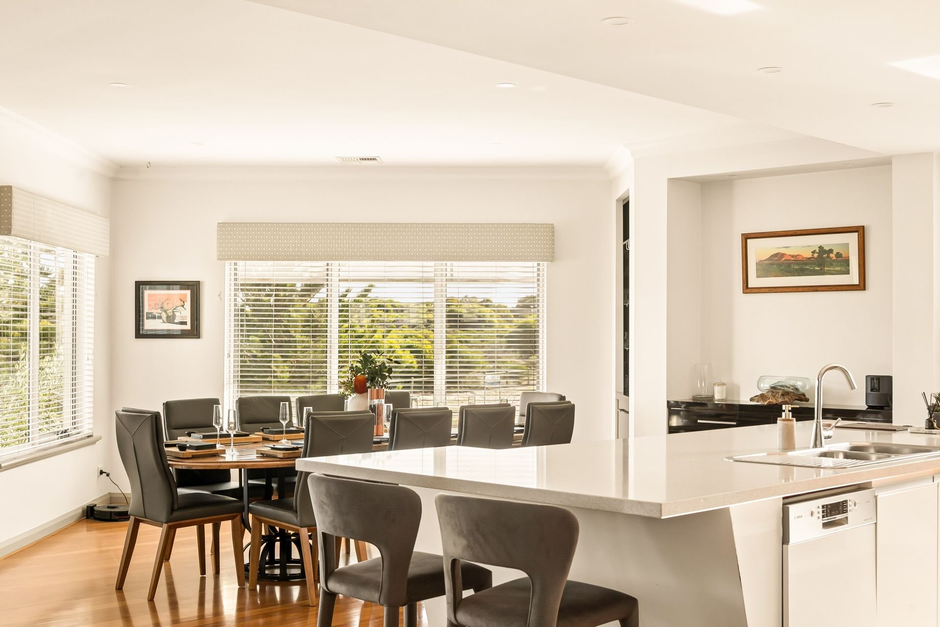Bright dining room with large windows, a round wooden table with black chairs, and a modern kitchen island with chairs. Wall art, plants, and kitchen sink are visible.