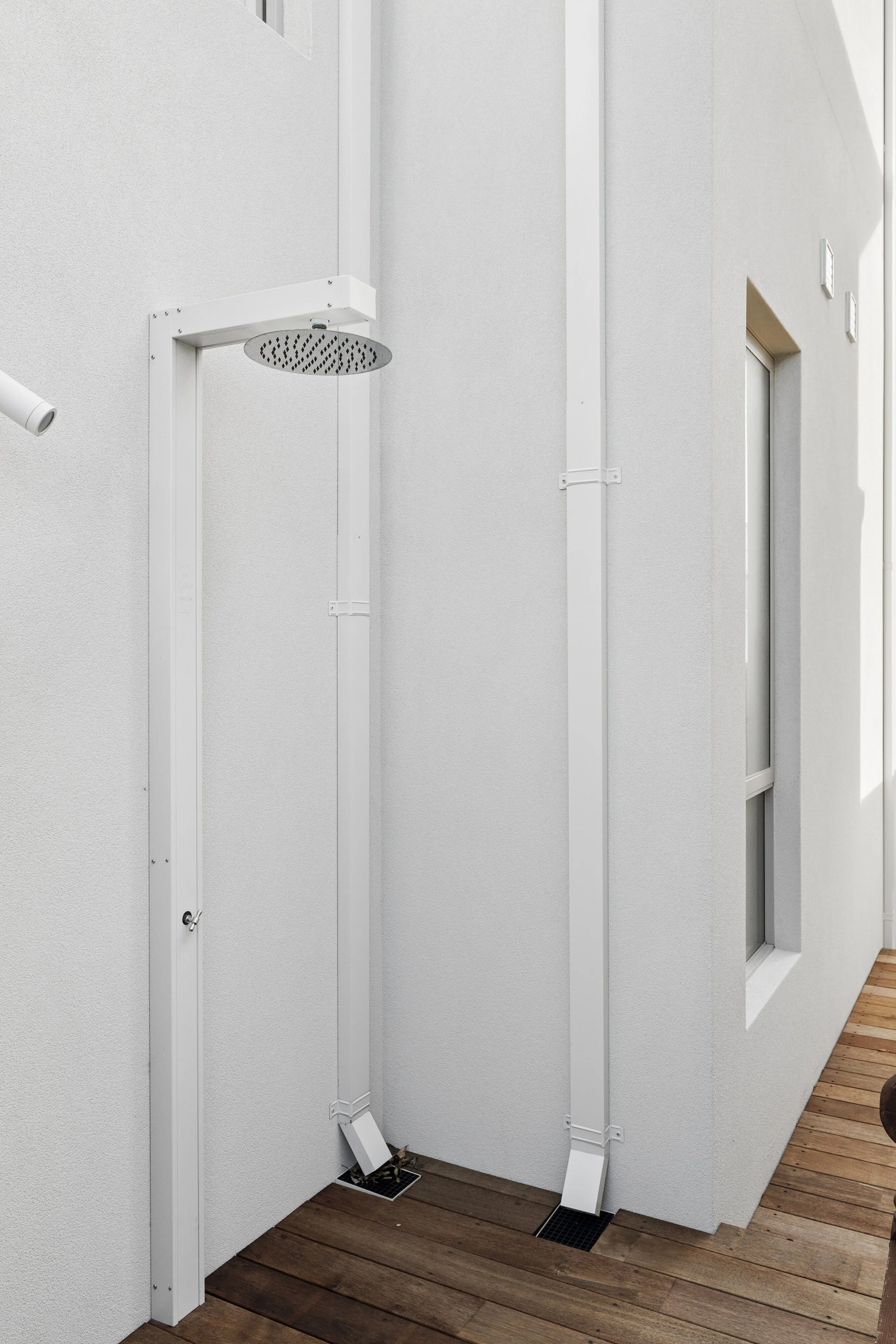 Outdoor shower on a wooden deck with white walls and a window in the background.
