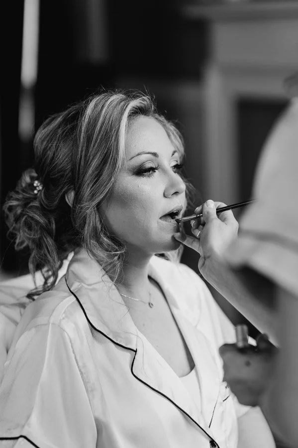 A bride with styled hair and light bridal makeup getting her lipstick applied by a makeup artist in a room.