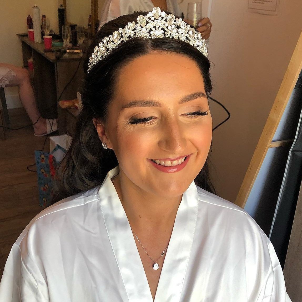 A bride smiling with eyes closed, wearing a pearl and crystal tiara, pearl earrings, a necklace, and a white satin robe.