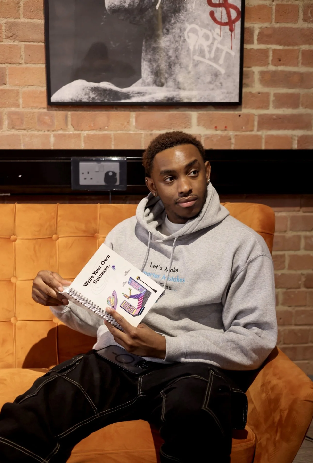 Young man sitting on an orange sofa with a brick wall background, holding a booklet titled "Write Your Own Universe".