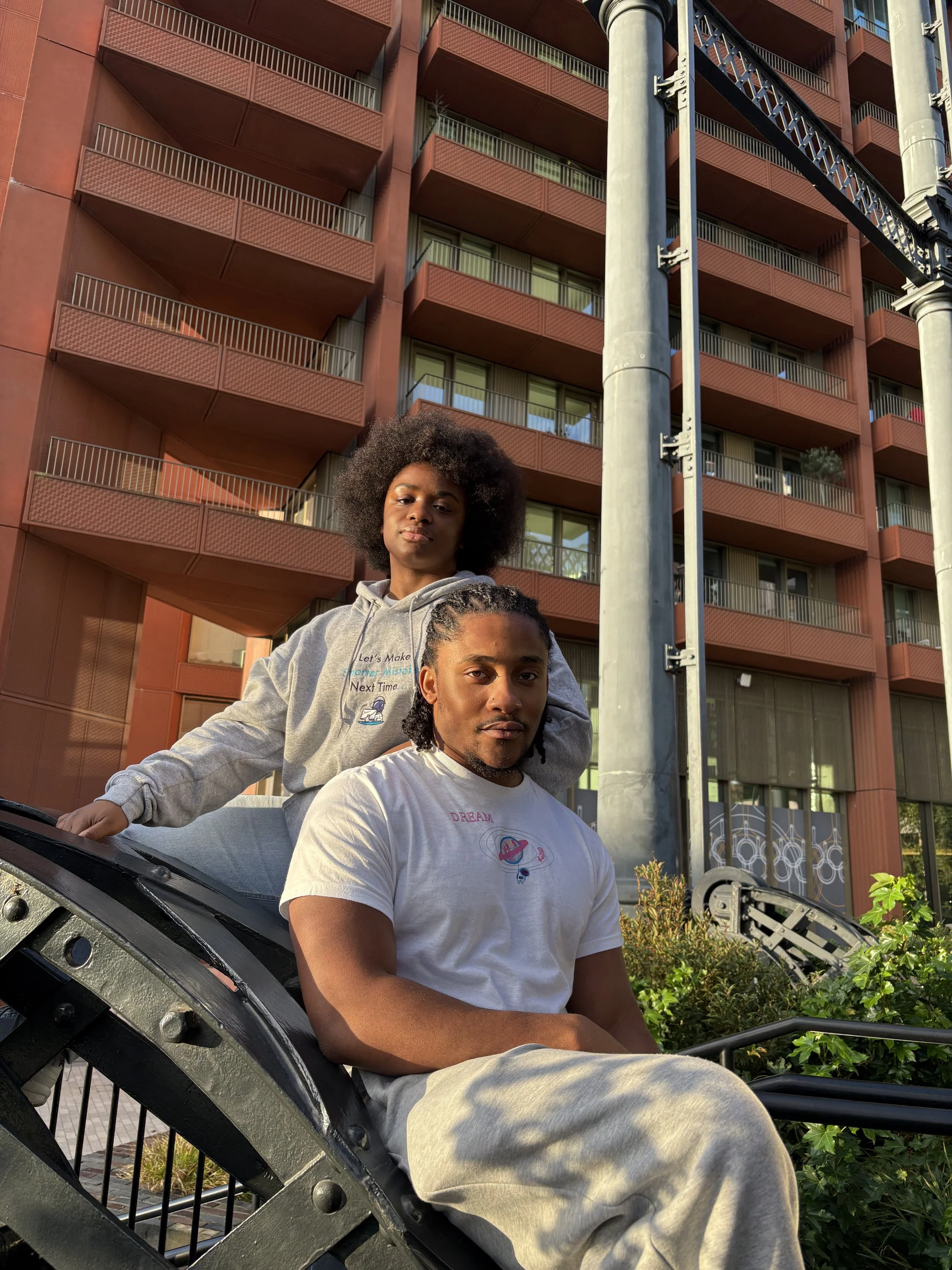 A young man and woman sitting on a park bench with a red apartment building and street lamp in the background.