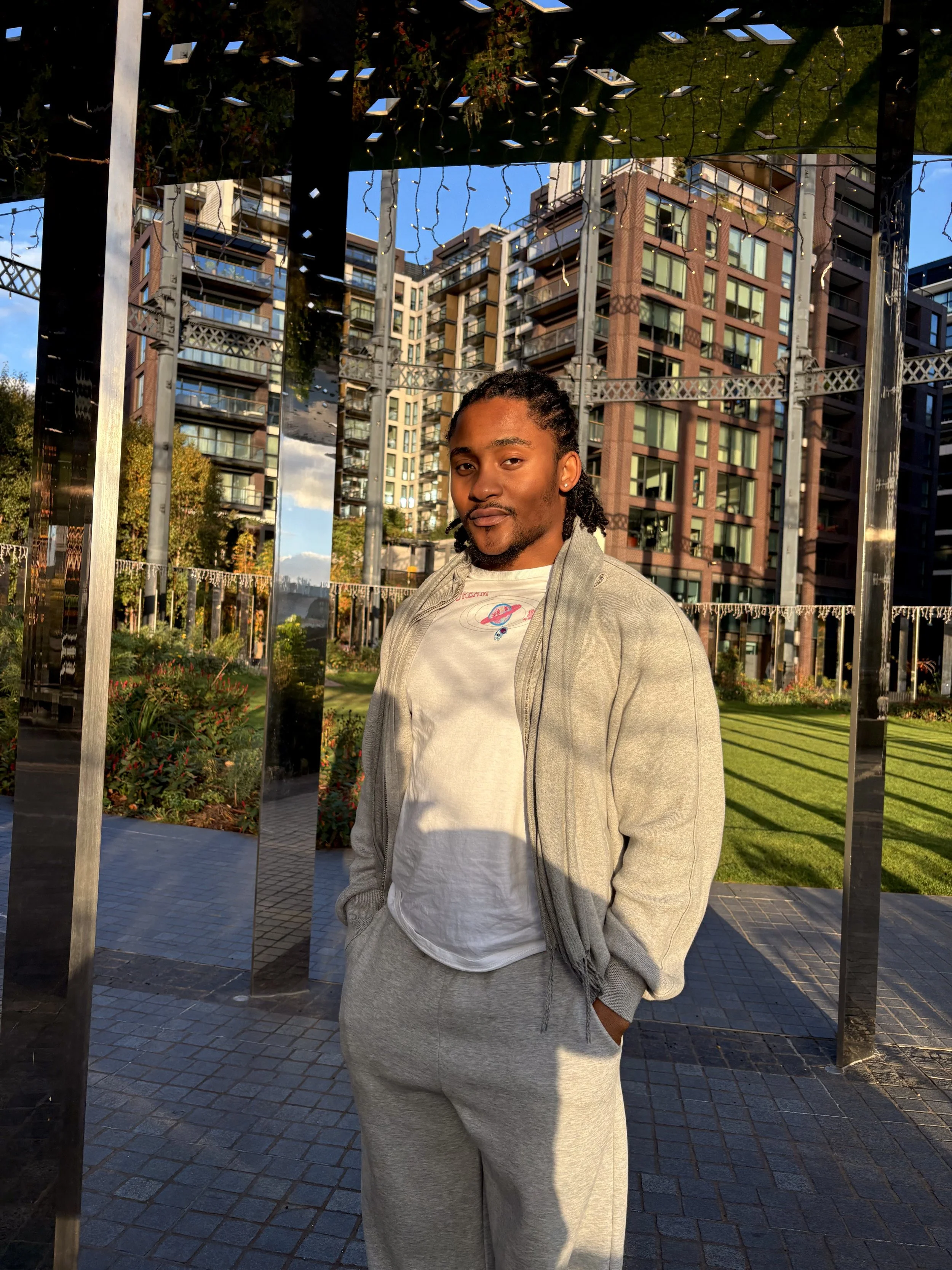 A young man standing outdoors in front of a reflective surface with high-rise apartment buildings in the background. He is wearing a light gray zip-up jacket over a white T-shirt and matching gray sweatpants. The man has his hands in his pockets and 