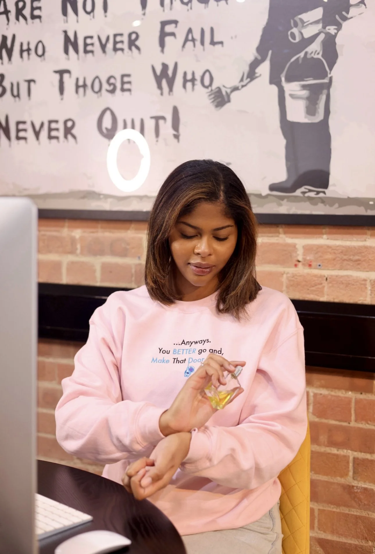 A young woman with shoulder-length hair in a pink sweatshirt, holding a small bottle of lotion or sanitizer and applying it to her wrist, sitting in a cafe or casual setting with a brick wall and a painting or poster in the background.