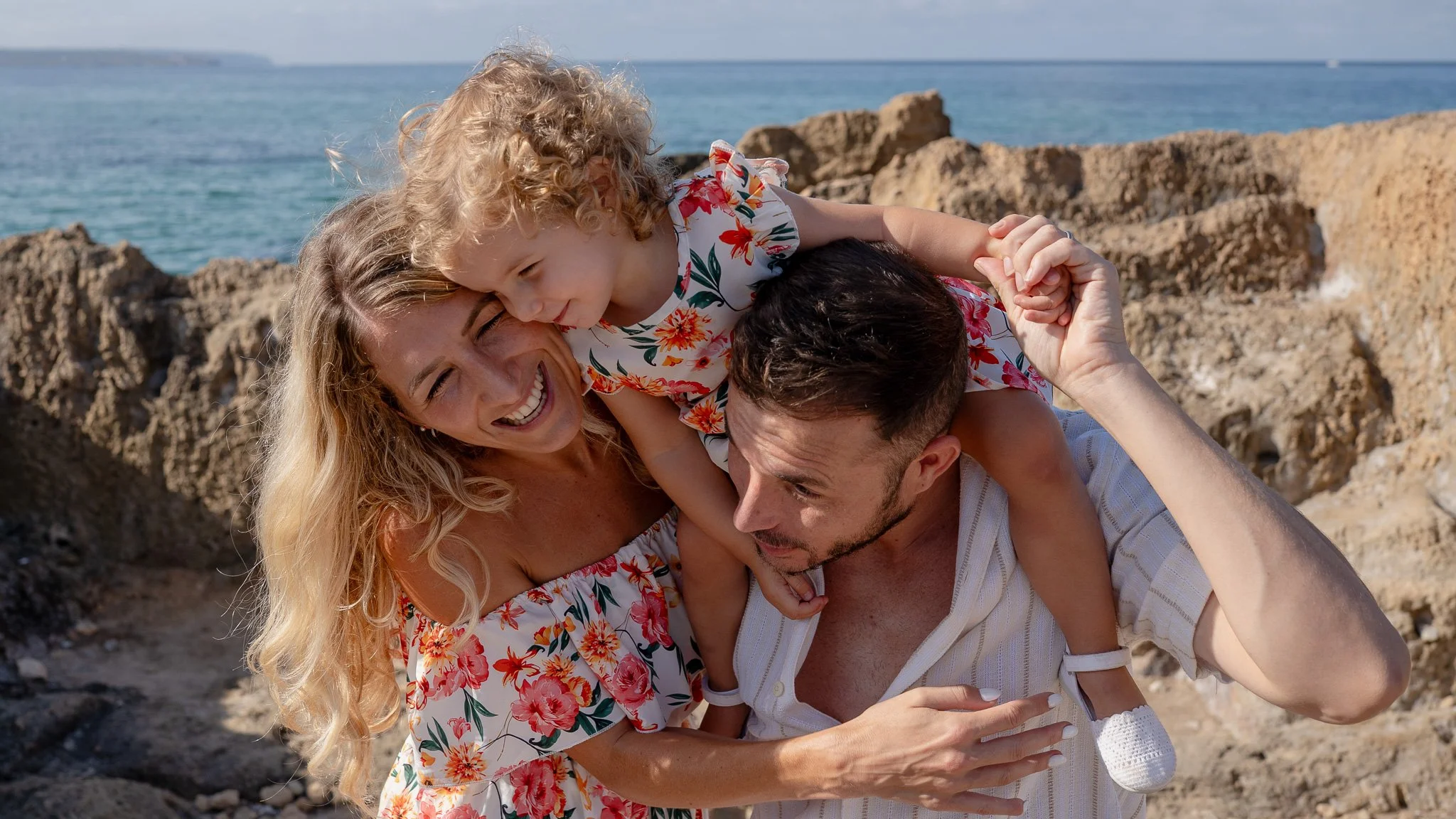 A family of three at the beach, with a woman, a man, and a young girl, all smiling and playfully interacting. The woman and girl are on the man's back, and they are surrounded by rocky terrain with the ocean and a clear sky in the background.