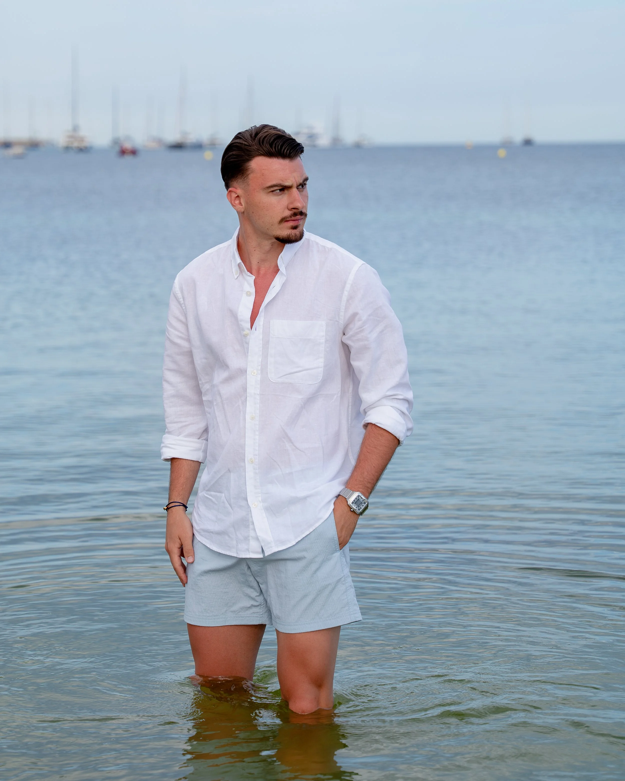 Natural male portrait photography in Mallorca, featuring a man standing in the sea wearing a white shirt and light blue trousers, looking thoughtfully to his left during a beach photoshoot.