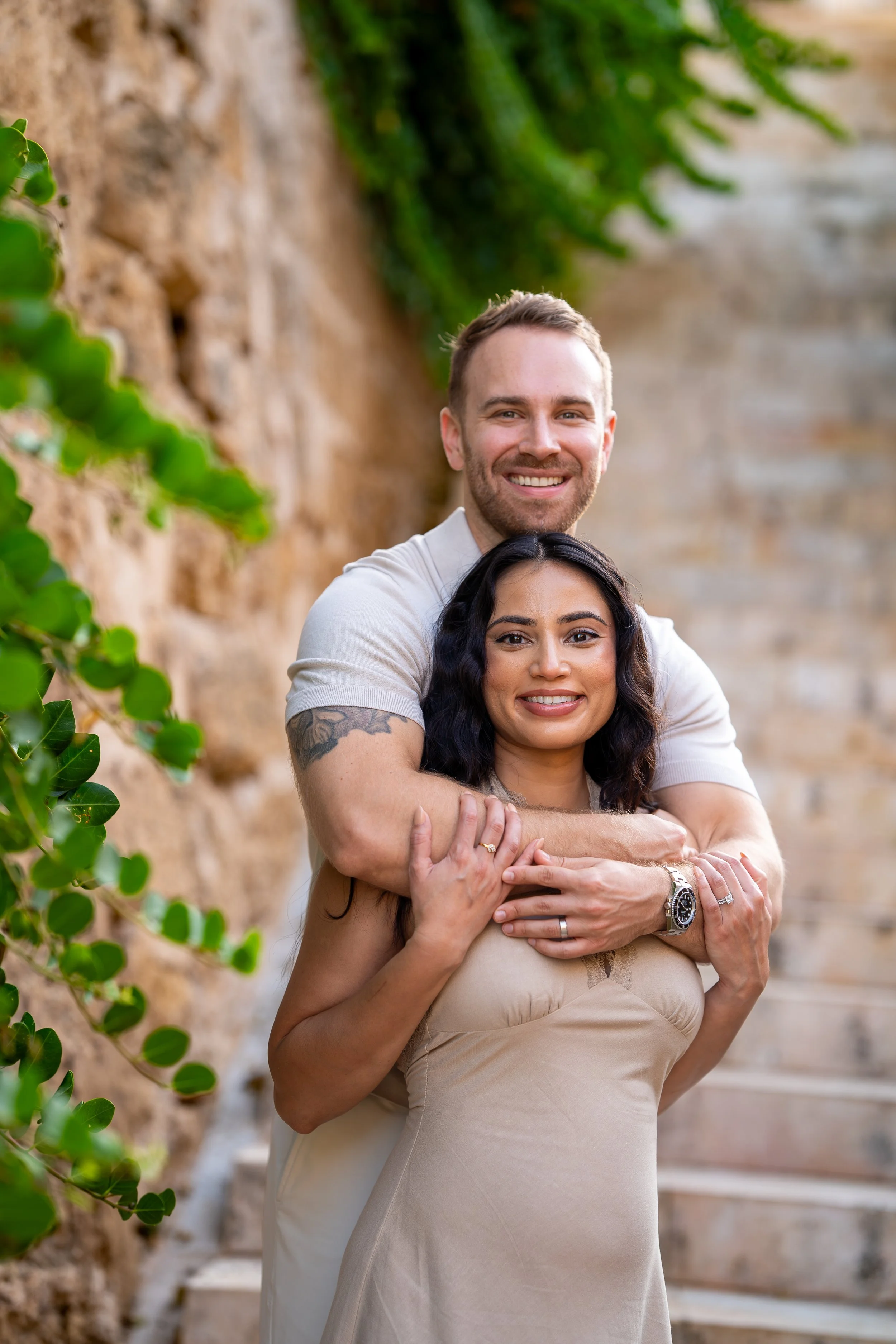 Romantic couple portrait in Mallorca, with a man standing behind his partner on a staircase, gently embracing her as they both look at the camera during a couples photoshoot.