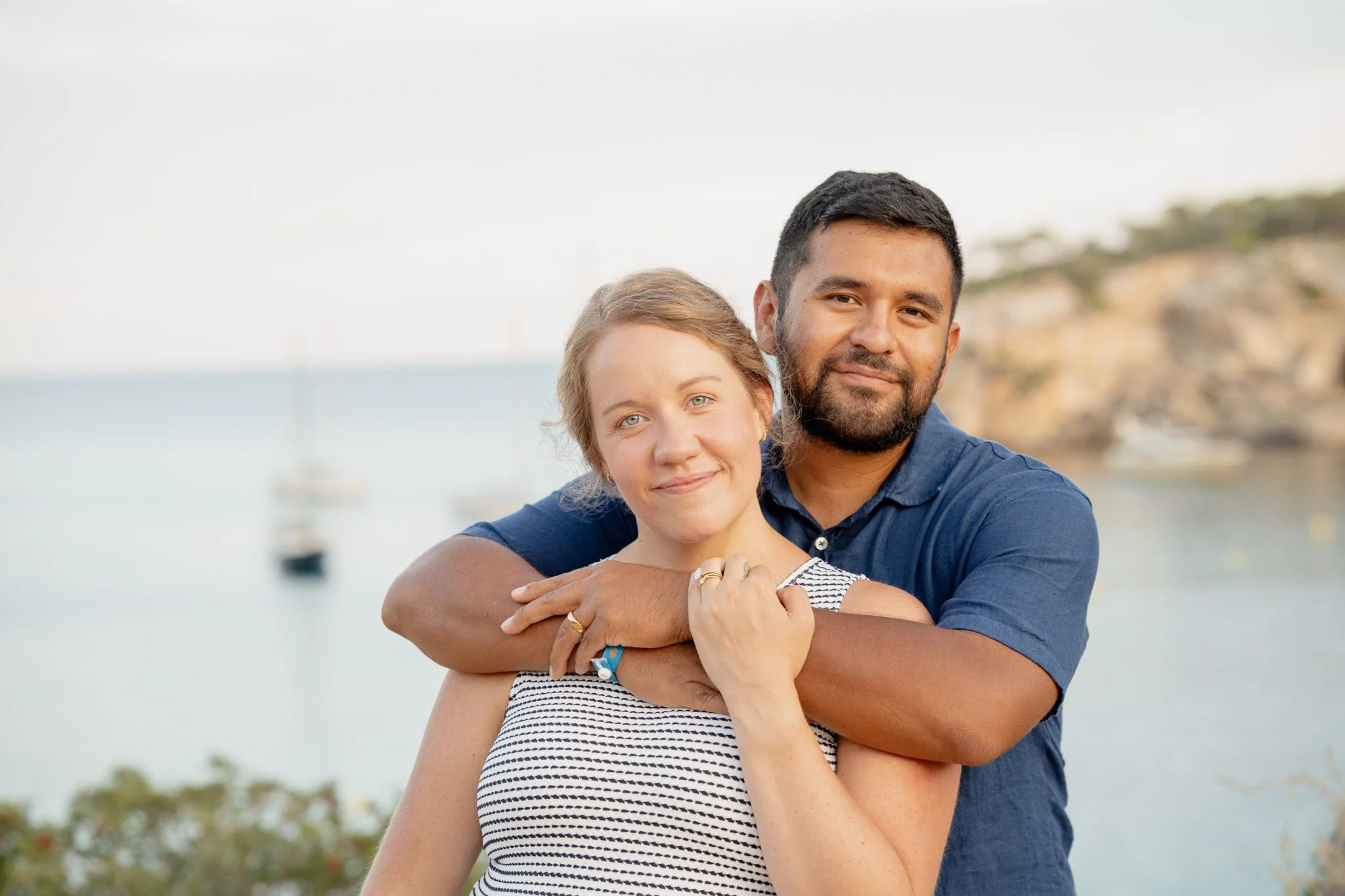 A couple embracing outdoors by the water, with boats and rocky cliffs in the background.