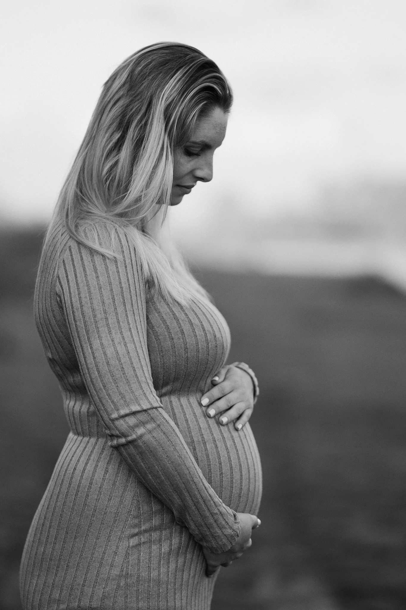A pregnant woman in a ribbed, long-sleeve dress standing outdoors with her eyes closed, holding her belly and gently touching her stomach.