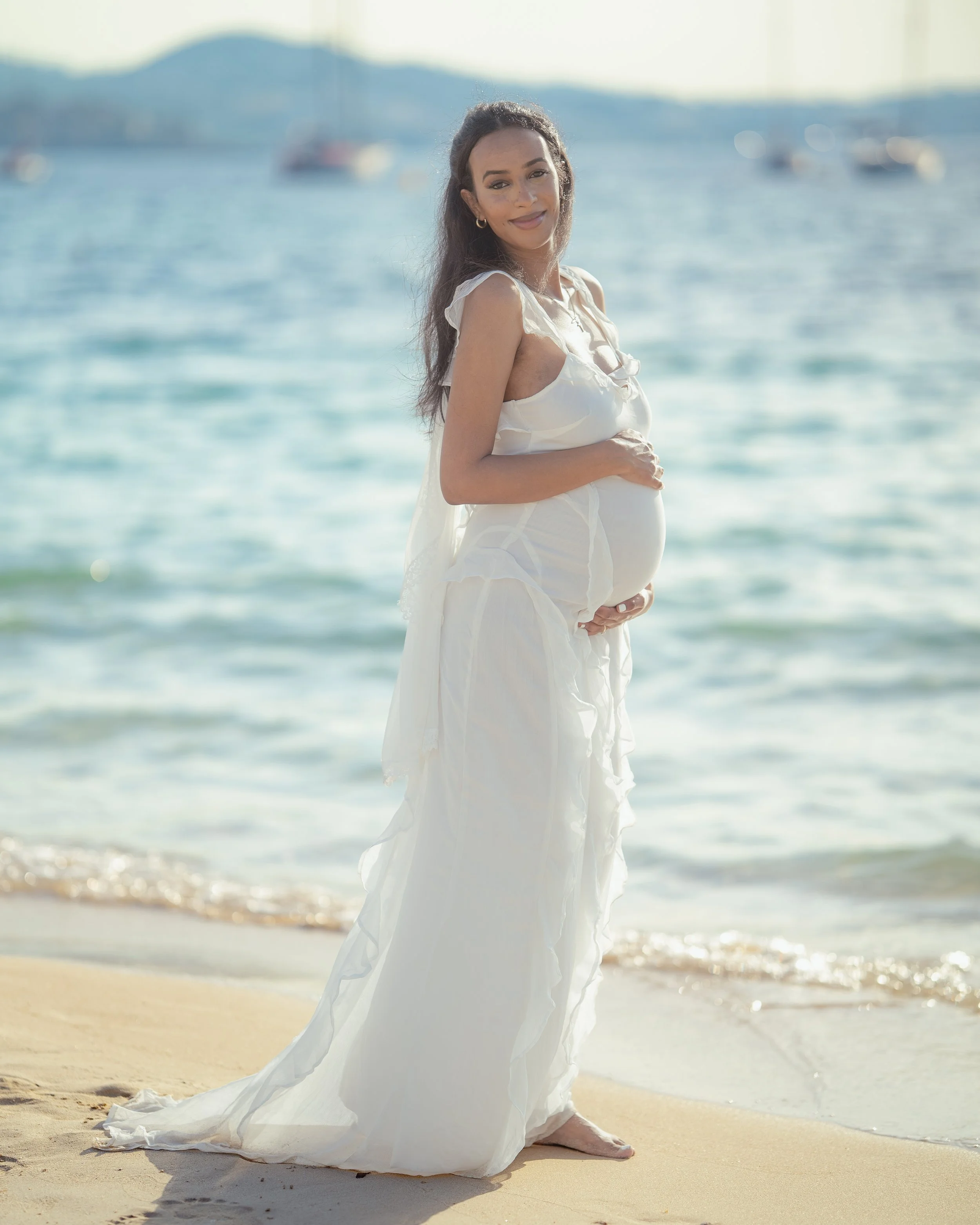 Zwangerschapsshoot op Mallorca met een aanstaande moeder in een zwierige witte jurk, die liefdevol haar zwangere buik vasthoudt met de turquoise zee als prachtig decor.