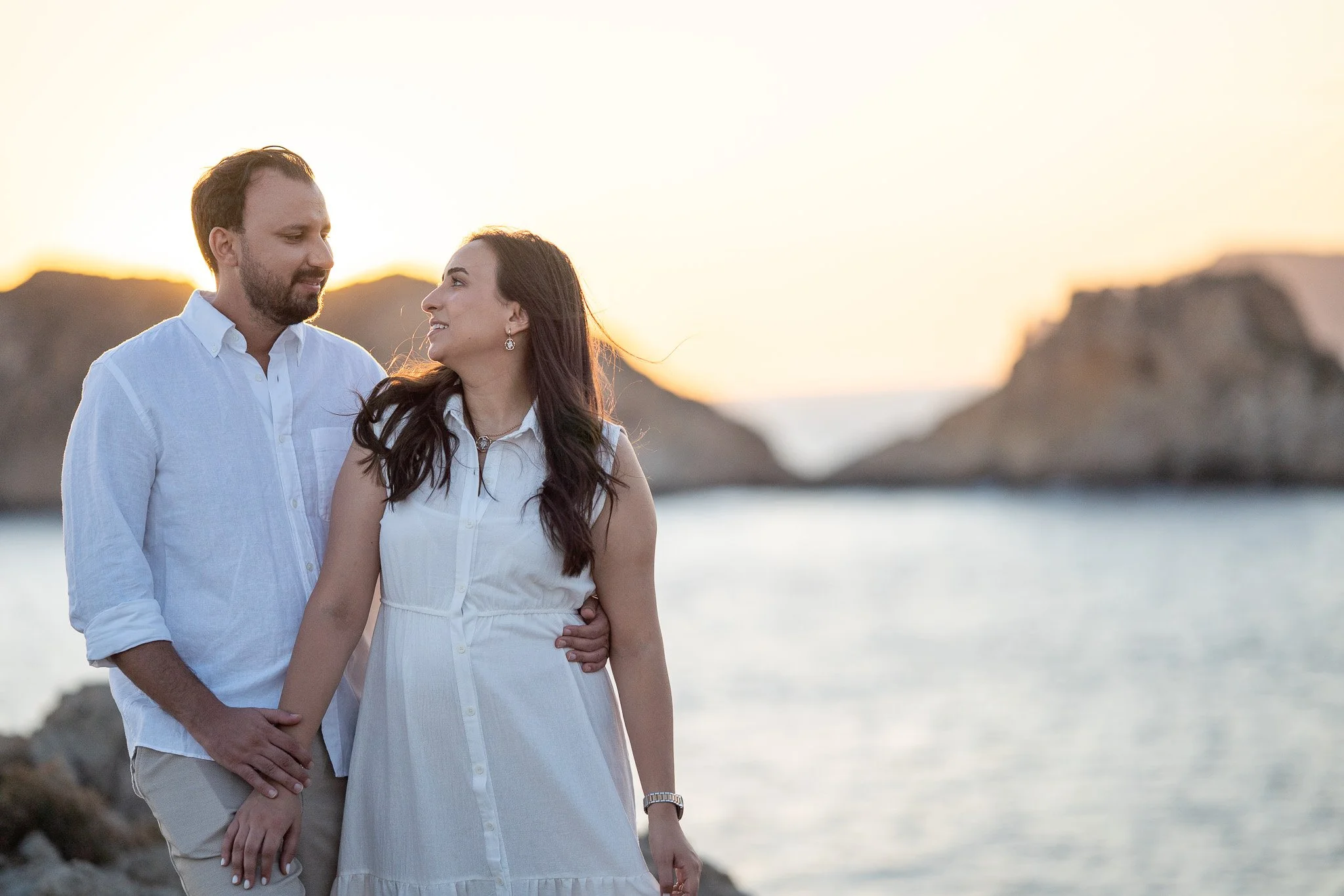 A couple stands by the water at sunset, looking into each other's eyes with a rocky landscape in the background in Mallorca