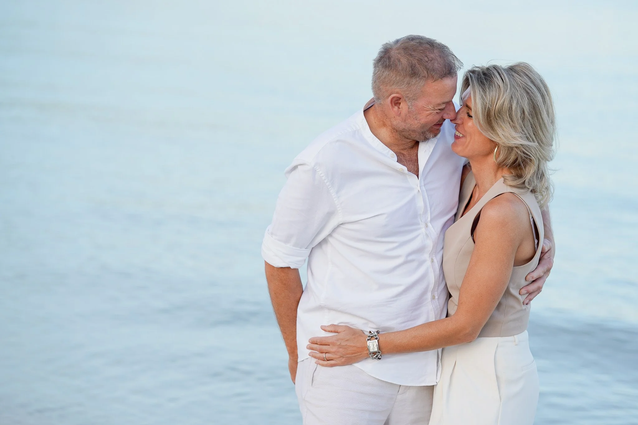A couple sharing a romantic moment near a body of water, smiling and touching foreheads.