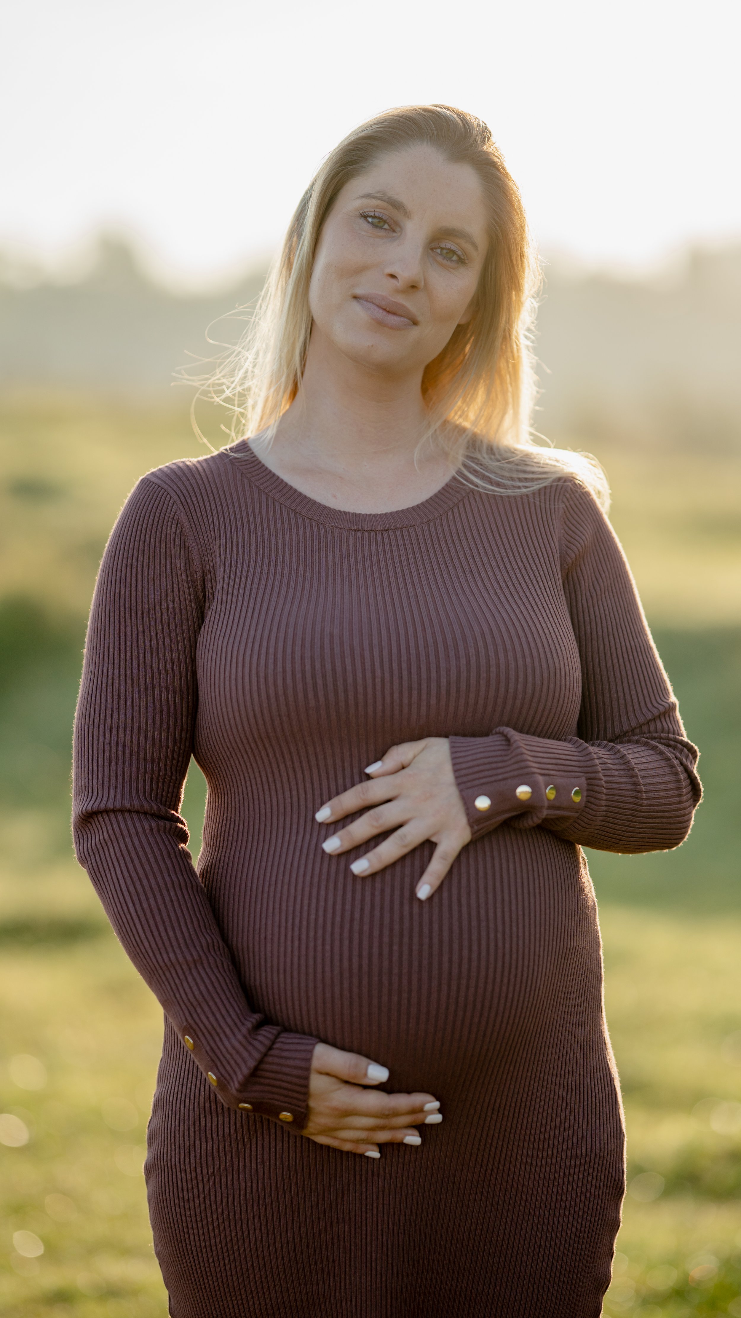 Elegante zwangerschapsshoot op Mallorca met een zwangere vrouw in een donkerrode bordeauxjurk, die liefdevol haar buik vasthoudt en recht in de camera kijkt tijdens een natuurlijke zwangerschapsfotosessie.