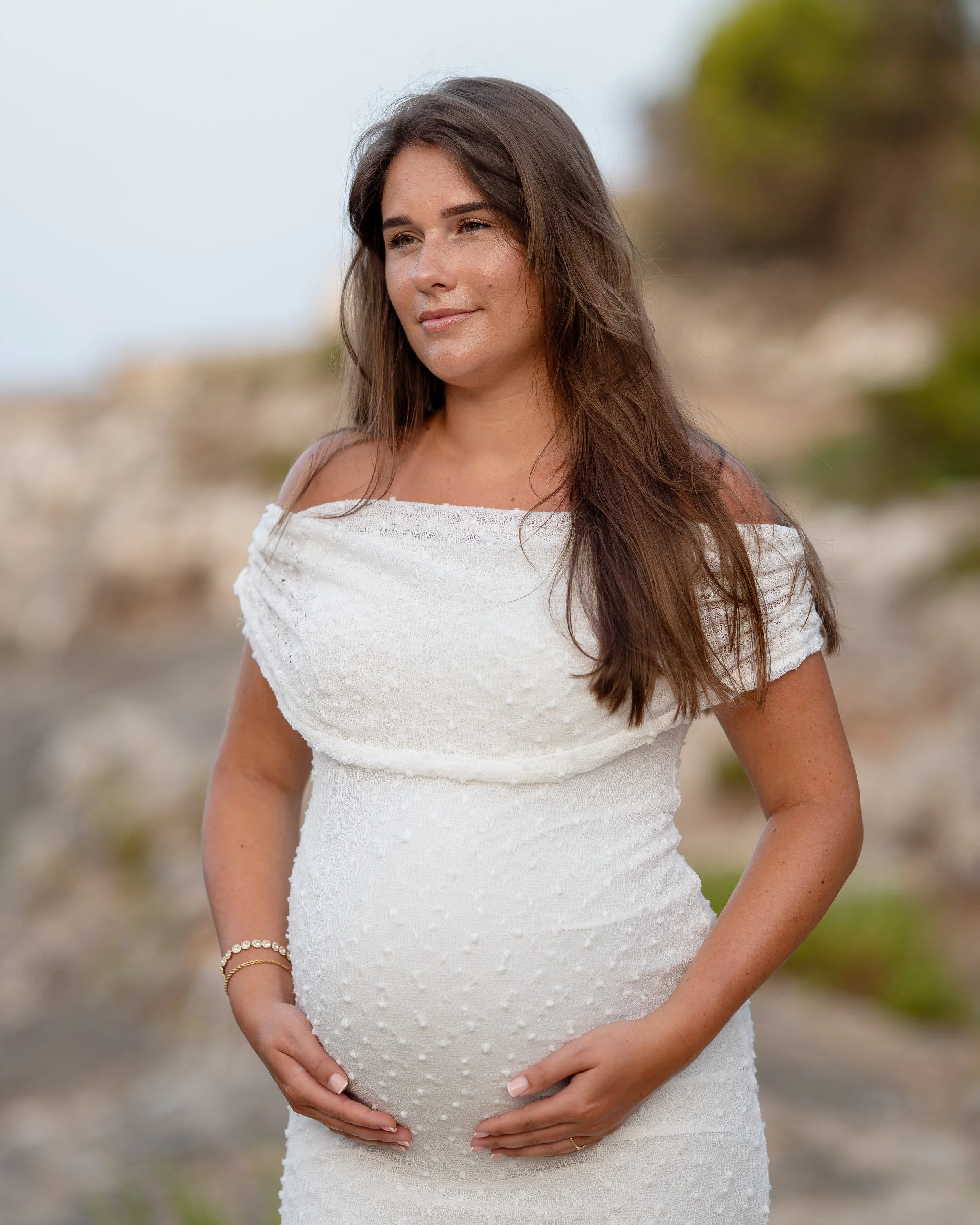 Zwangerschapsshoot op Mallorca met een zwangere vrouw in een prachtige witte jurk die tijdens een elegante, natuurlijke portretsessie dromerig in de verte kijkt.