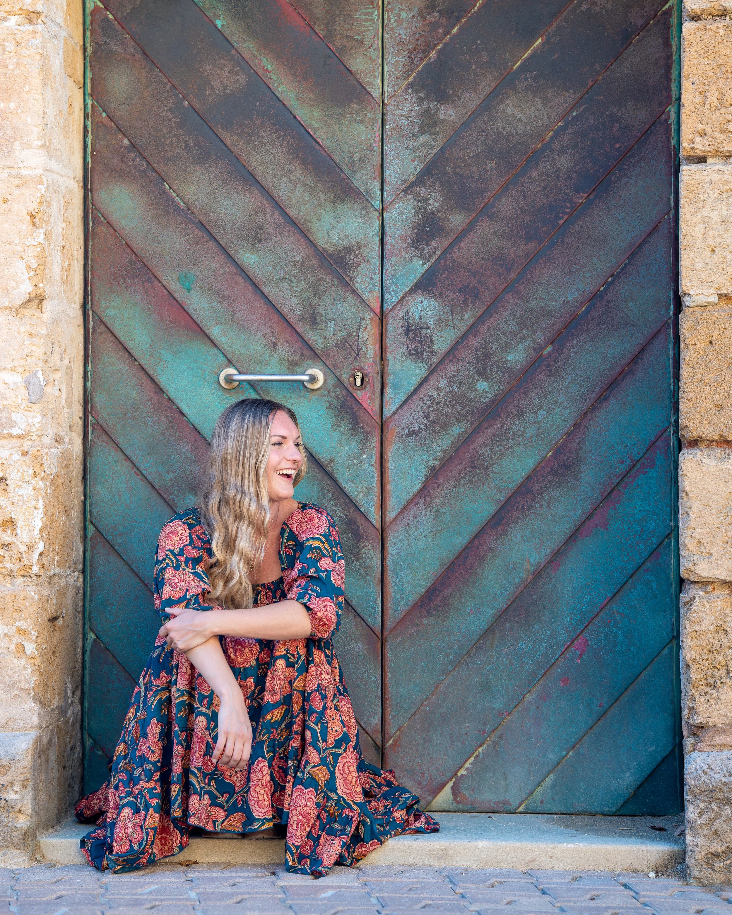 Smiling woman posing in the streets of Palma de Mallorca, crouching with her back against a doorway and looking to her left during a natural urban portrait photoshoot.