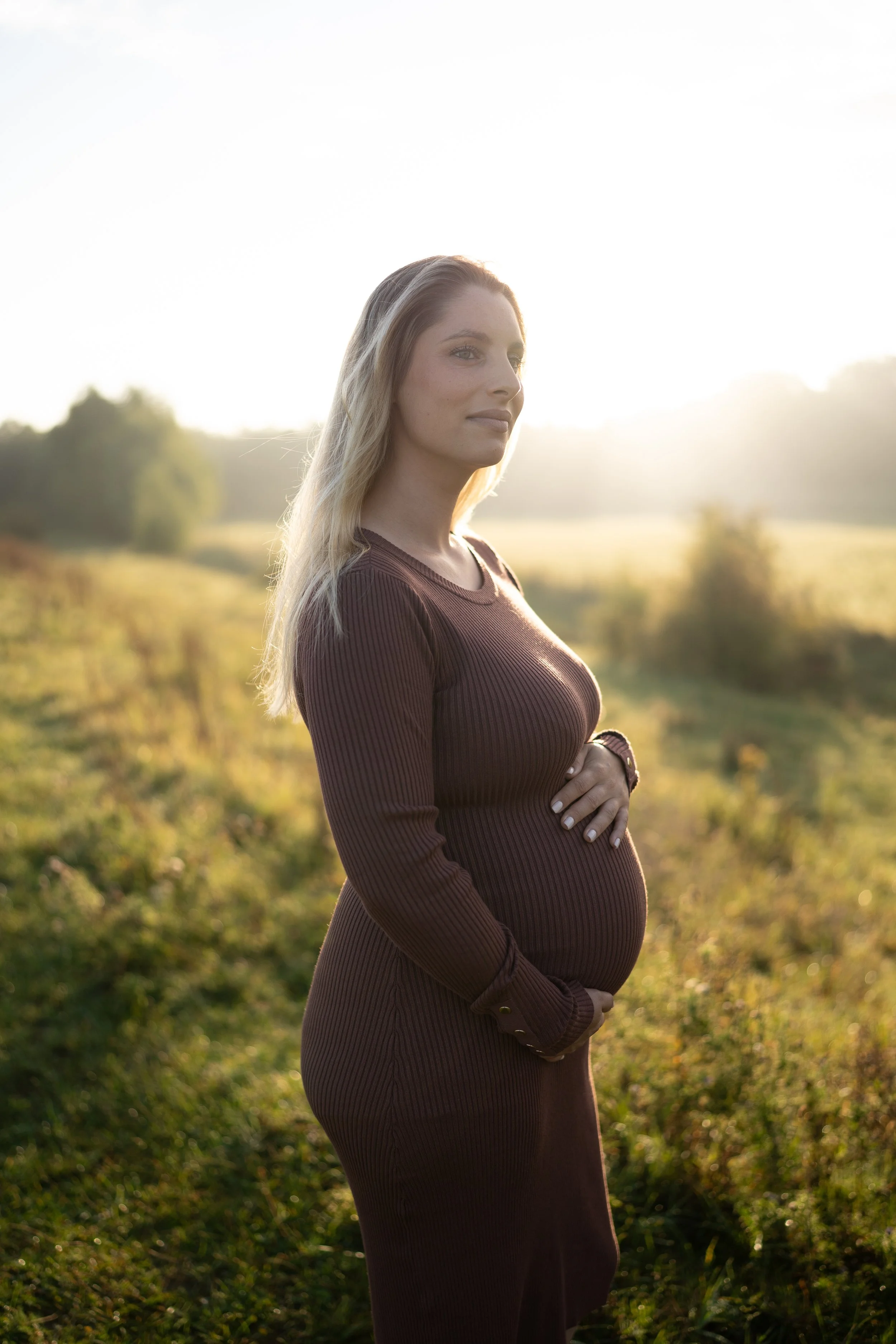 A pregnant woman in a brown long-sleeve dress standing outdoors in a field during sunset in the Netherlands.