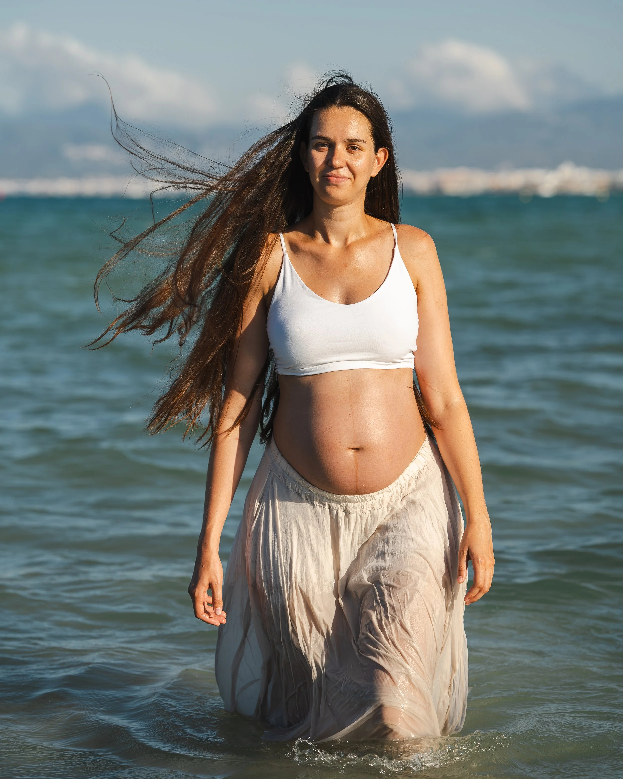 Dromerige zwangerschapsshoot op Mallorca met een zwangere vrouw die in zee staat, terwijl de zeebries zacht door haar lange haren waait tijdens een sfeervolle zwangerschapsfotoshoot op het strand.