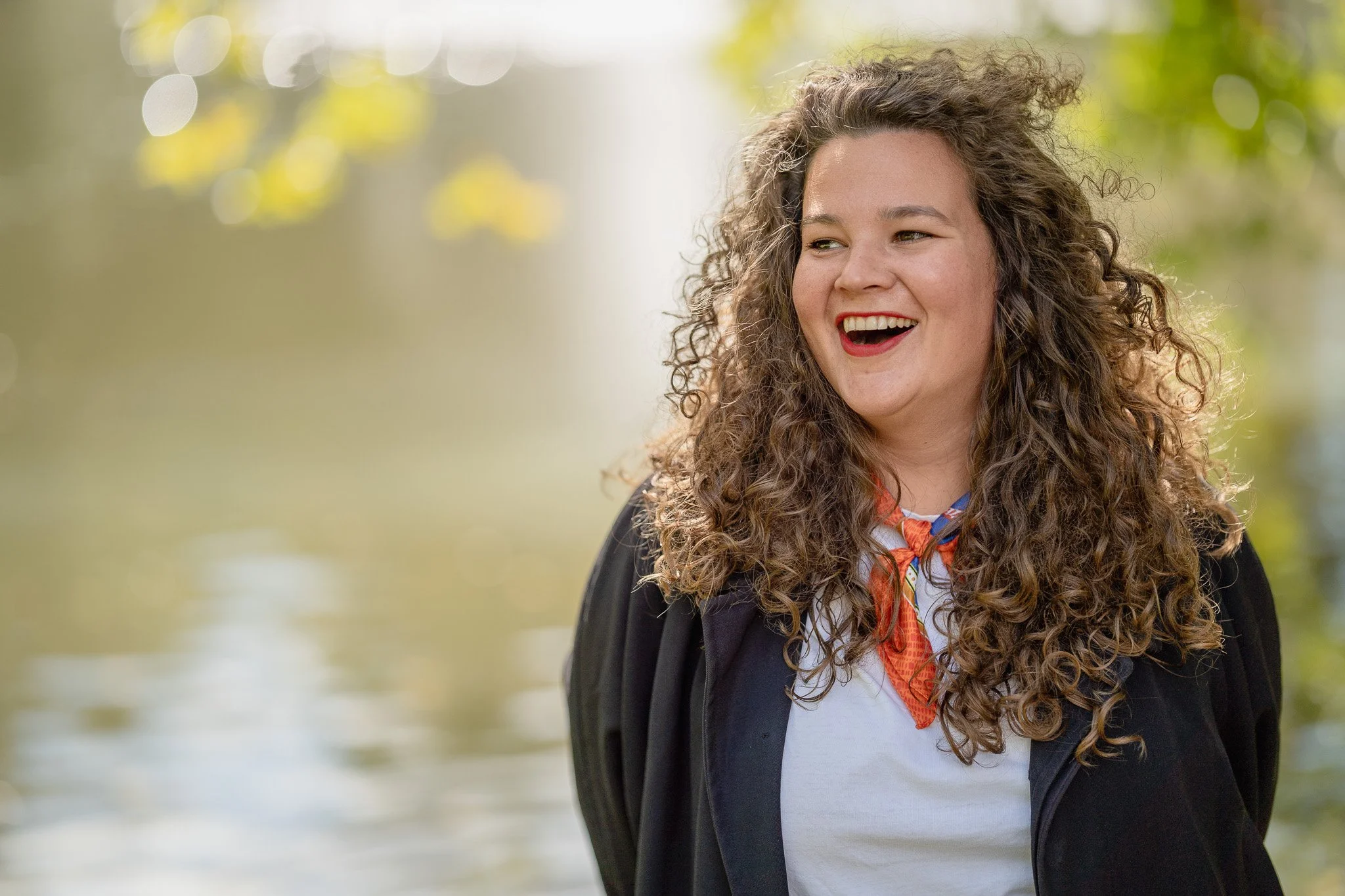 A smiling woman with long curly hair, wearing a white shirt and an orange neckerchief, outdoors with blurred green foliage and water in the background.