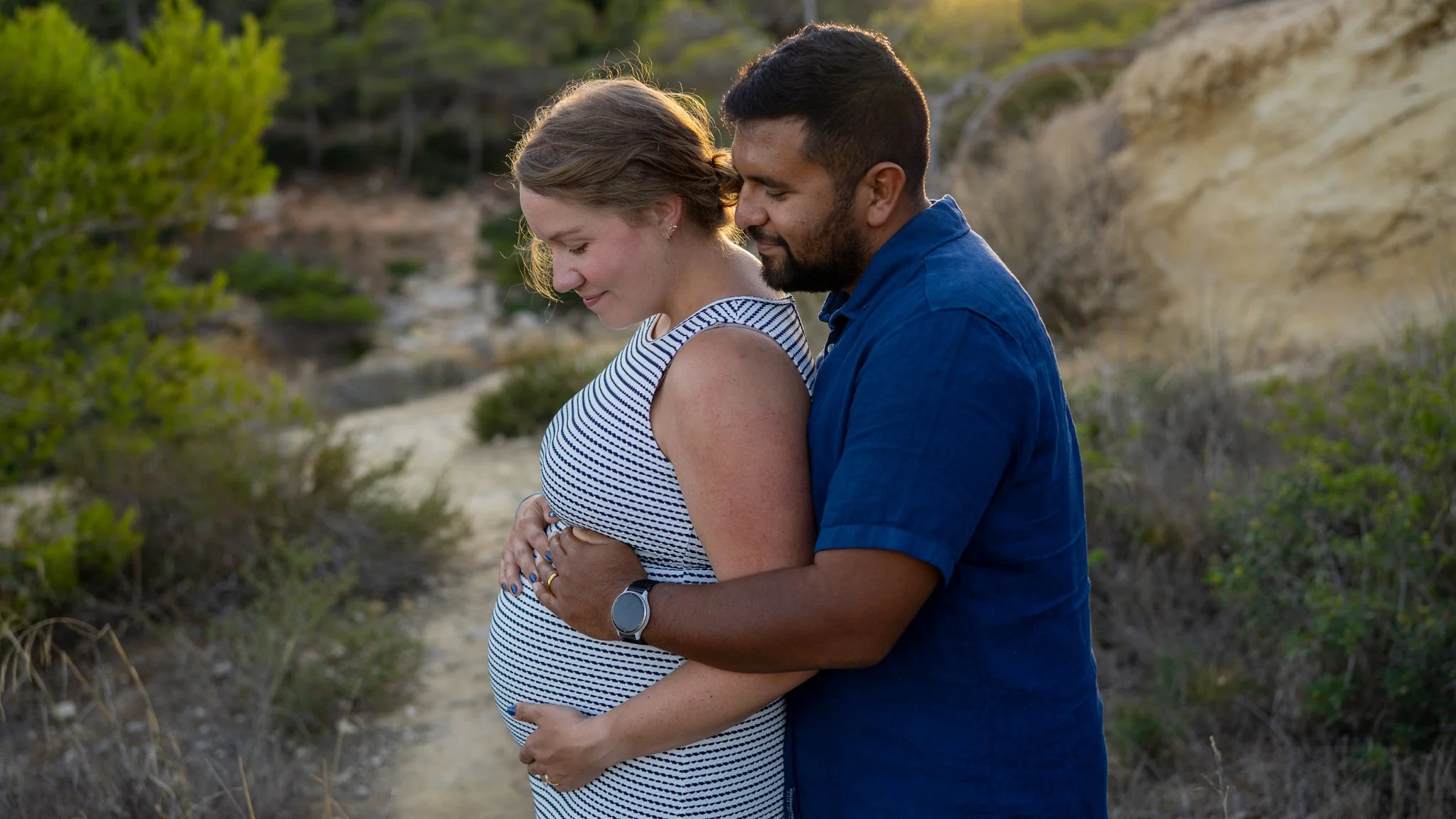 A pregnant woman and a man standing close together on a dirt path outdoors, with trees and hills in the background, during sunset, sharing an intimate moment.