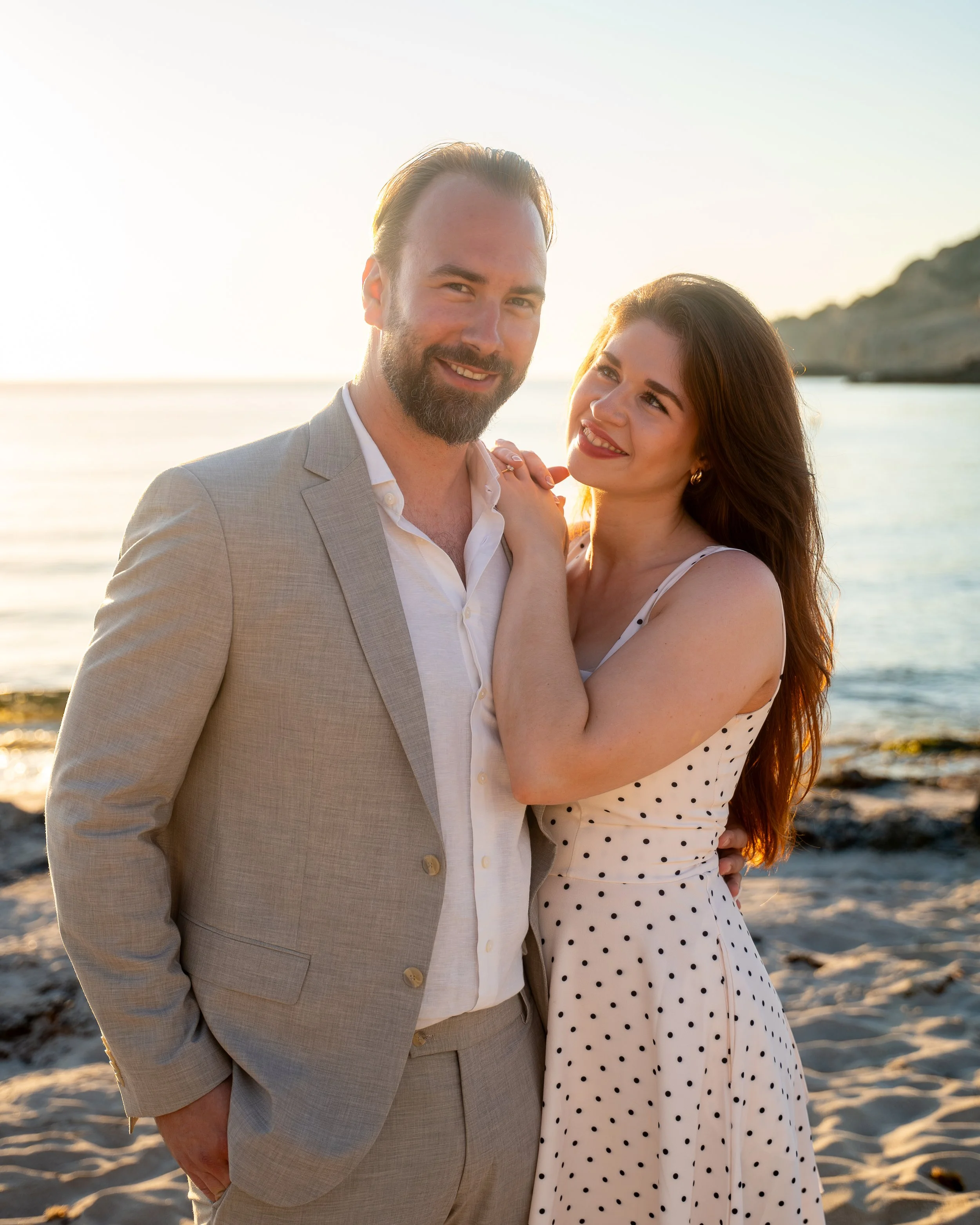 Romantische koppelfotoshoot op Mallorca tijdens zonsopgang op het strand, waarbij een vrouw haar man liefdevol aankijkt met haar handen op zijn schouders.