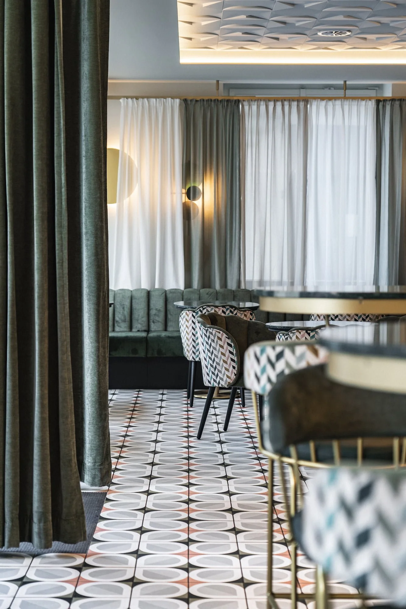 Stilvoller Innenraum des Rio Café & Cocktailbar in Dornbirn mit gemustertem Fliesenboden, grünen Vorhängen, Polsterstühlen und grüner Couch.