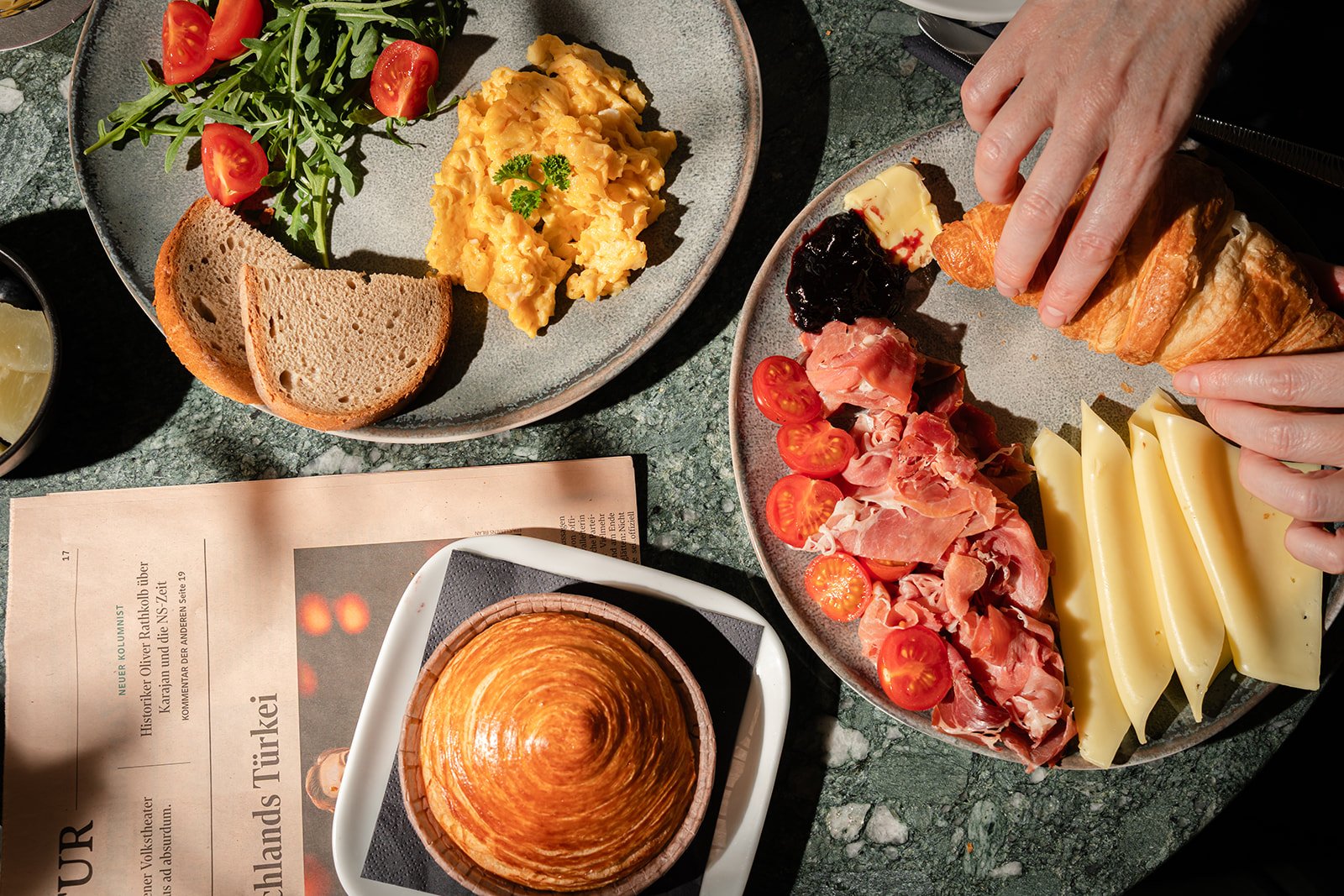 Frühstücksteller mit Tomaten, Rucola, Rührei, Vollkornbrot, und verschiedene Wurst- und Käsesorten, sowie ein gekochtes Ei in einer Schale.