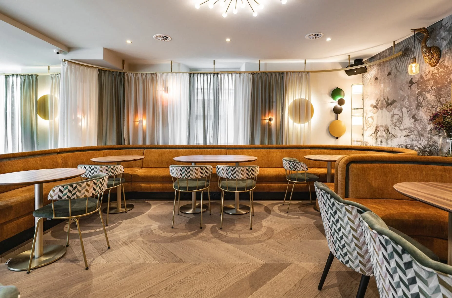 Moderner, eleganter Innenraum des Rio Café & Cocktailbar in Dornbirn mit Rundbänken, Wandlampen, Vorhängen mit Lichtakzenten und Vogel-Wandkunst.