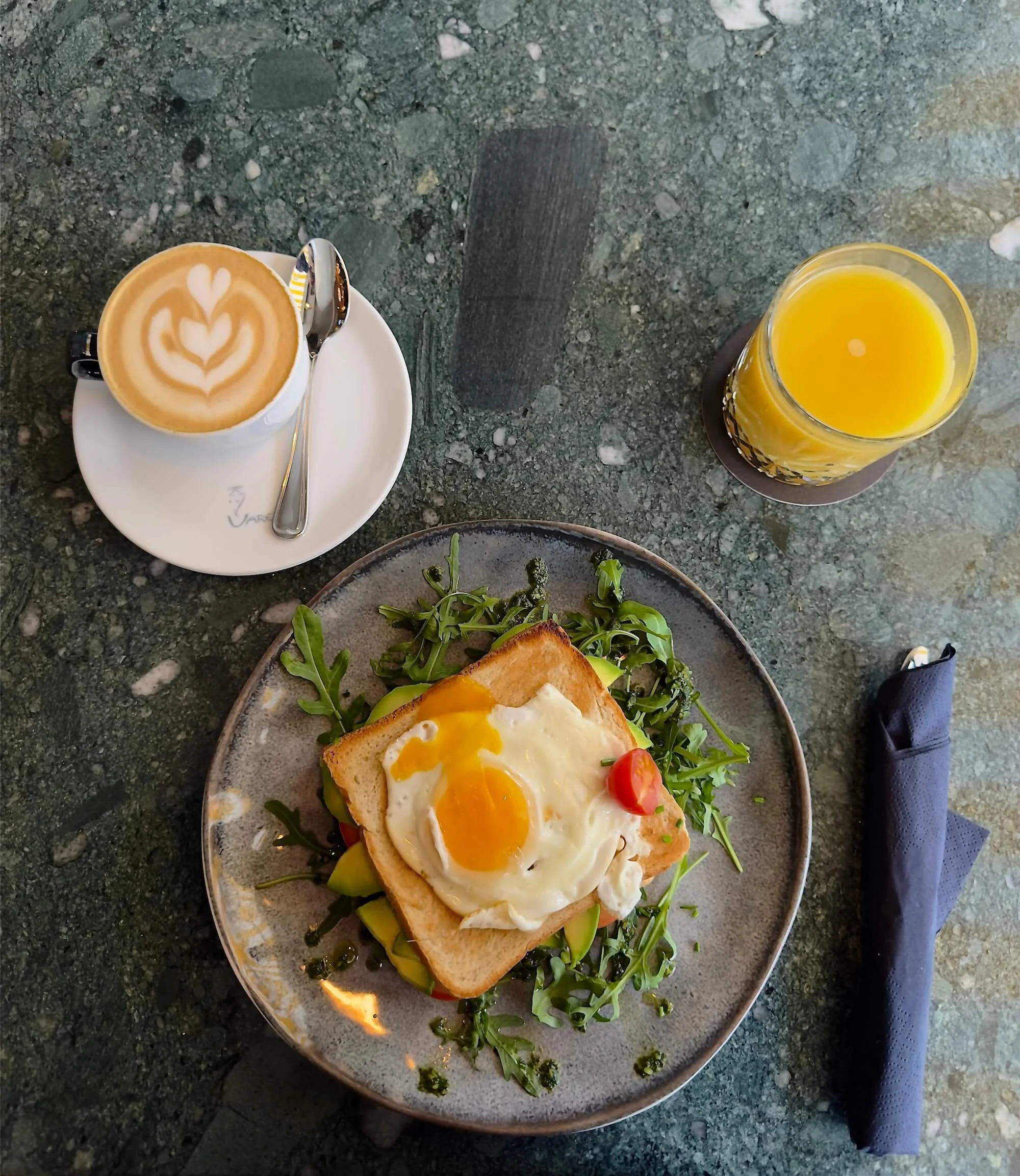 Ein Teller mit einem Toast, belegt mit einem Spiegelei, Salat und Tomaten, daneben ein Glas Orangensaft und eine Kaffee-Tasse mit Latte Art.