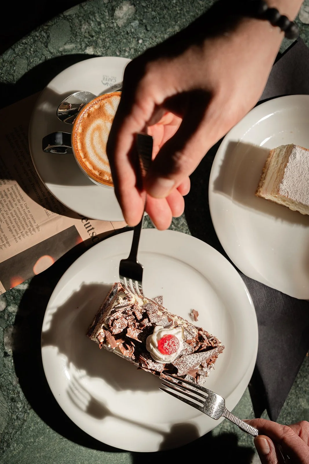Ein Stück Schokoladenkuchen mit Sahne und Kirsche auf einem weißen Teller, eine Tasse Kaffee mit Milchschaum, eine Person nimmt mit einer Gabel am Kuchen, auf einem grünen Marmor-Tisch