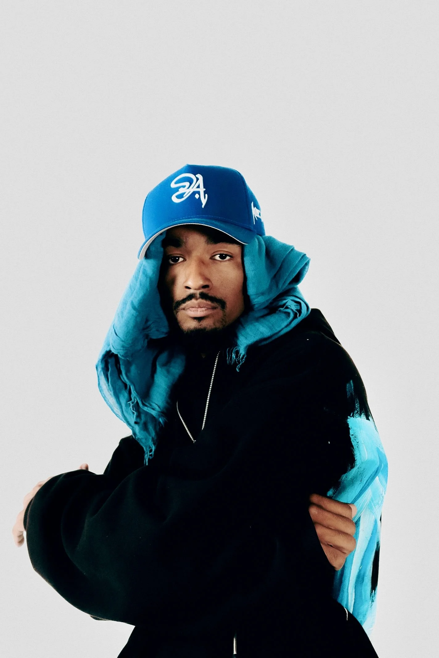 A man wearing a blue baseball cap, black jacket with blue and white designs, and a blue hoodie draped around his neck, posing against a plain light gray background.