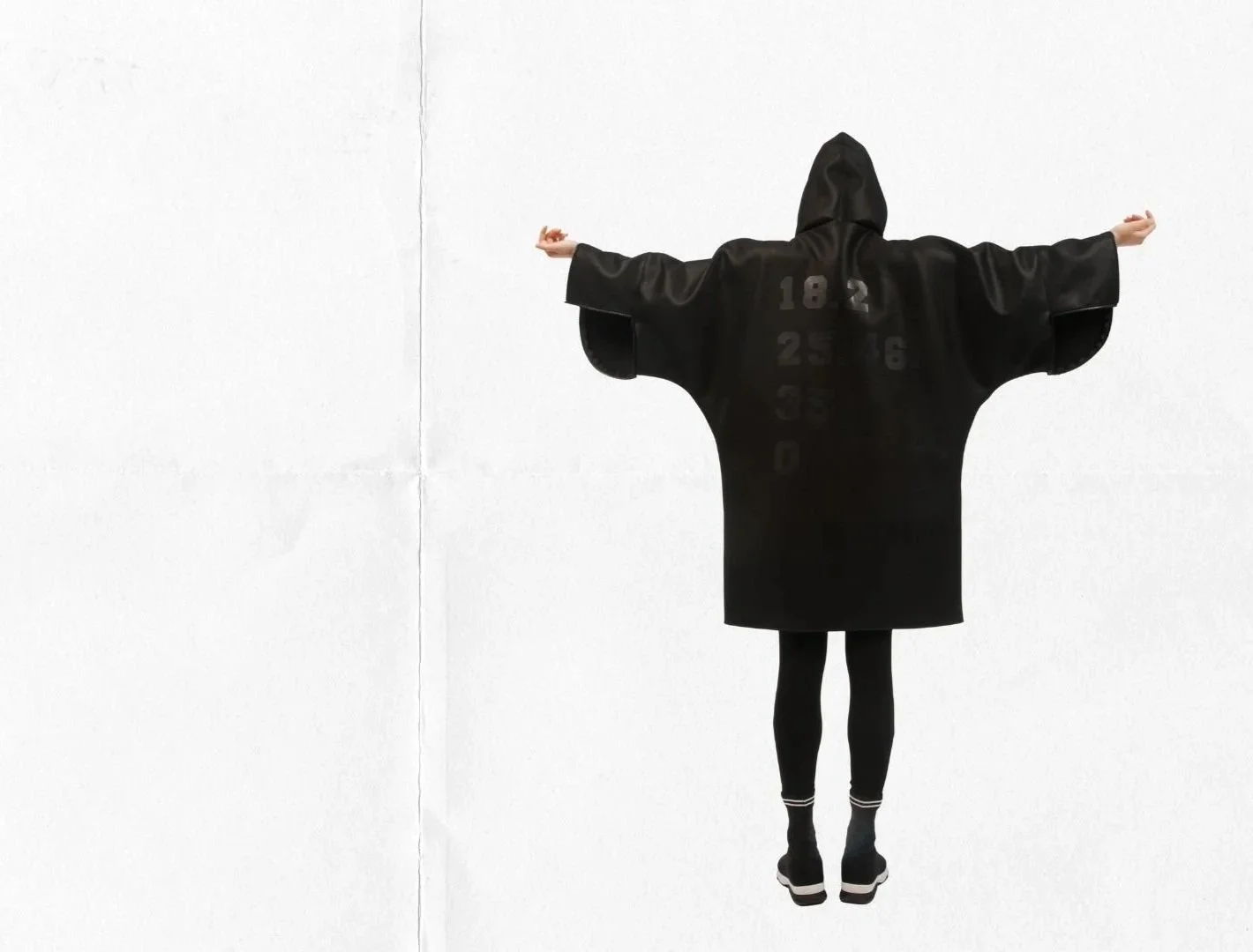 Person wearing a black hooded robe with numbers printed on the back, standing with arms outstretched against a white wall.