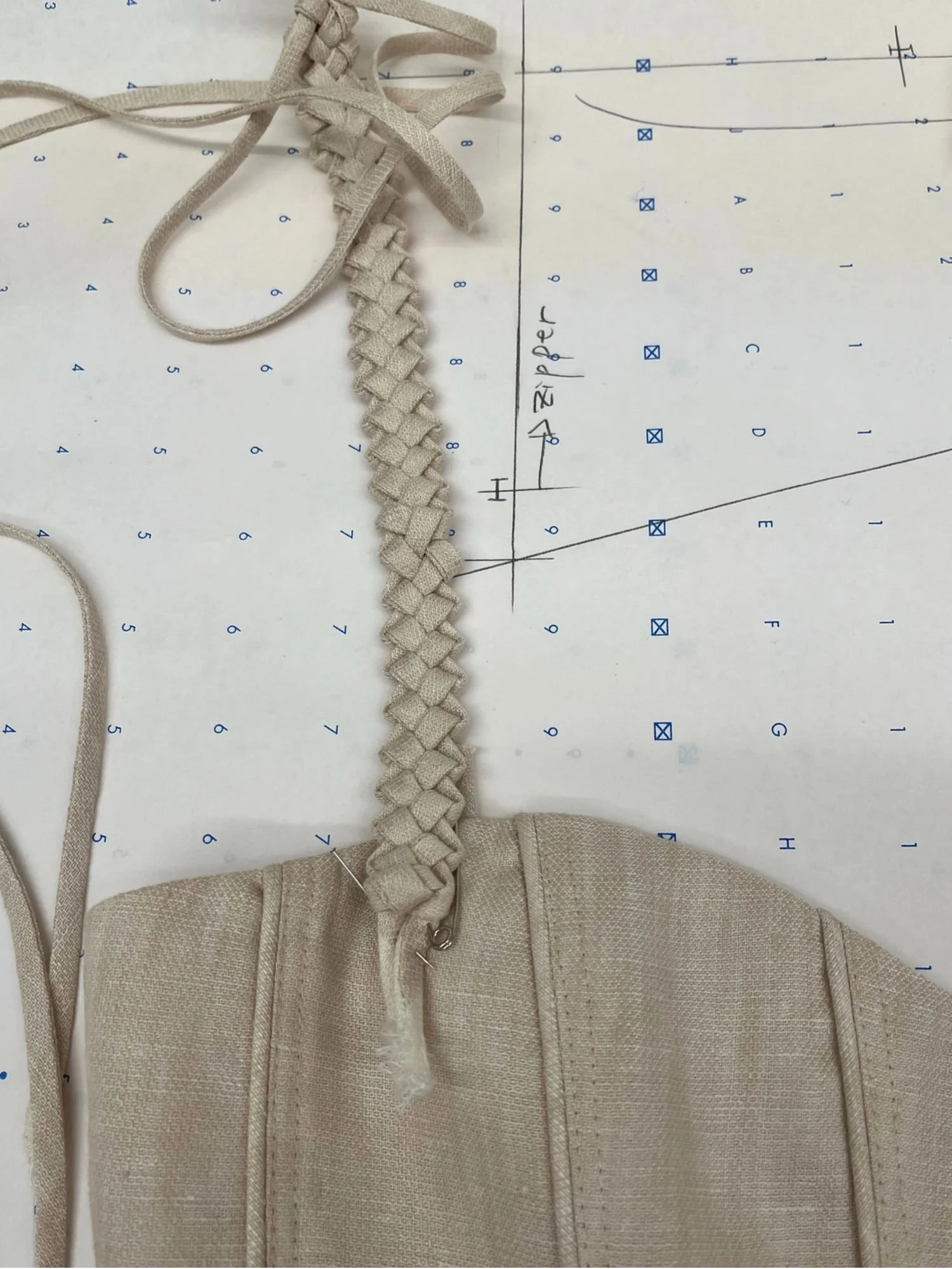 Beige fabric mask with an attached beige braided cord, placed on a double-sided grid paper with handwriting and markings.
