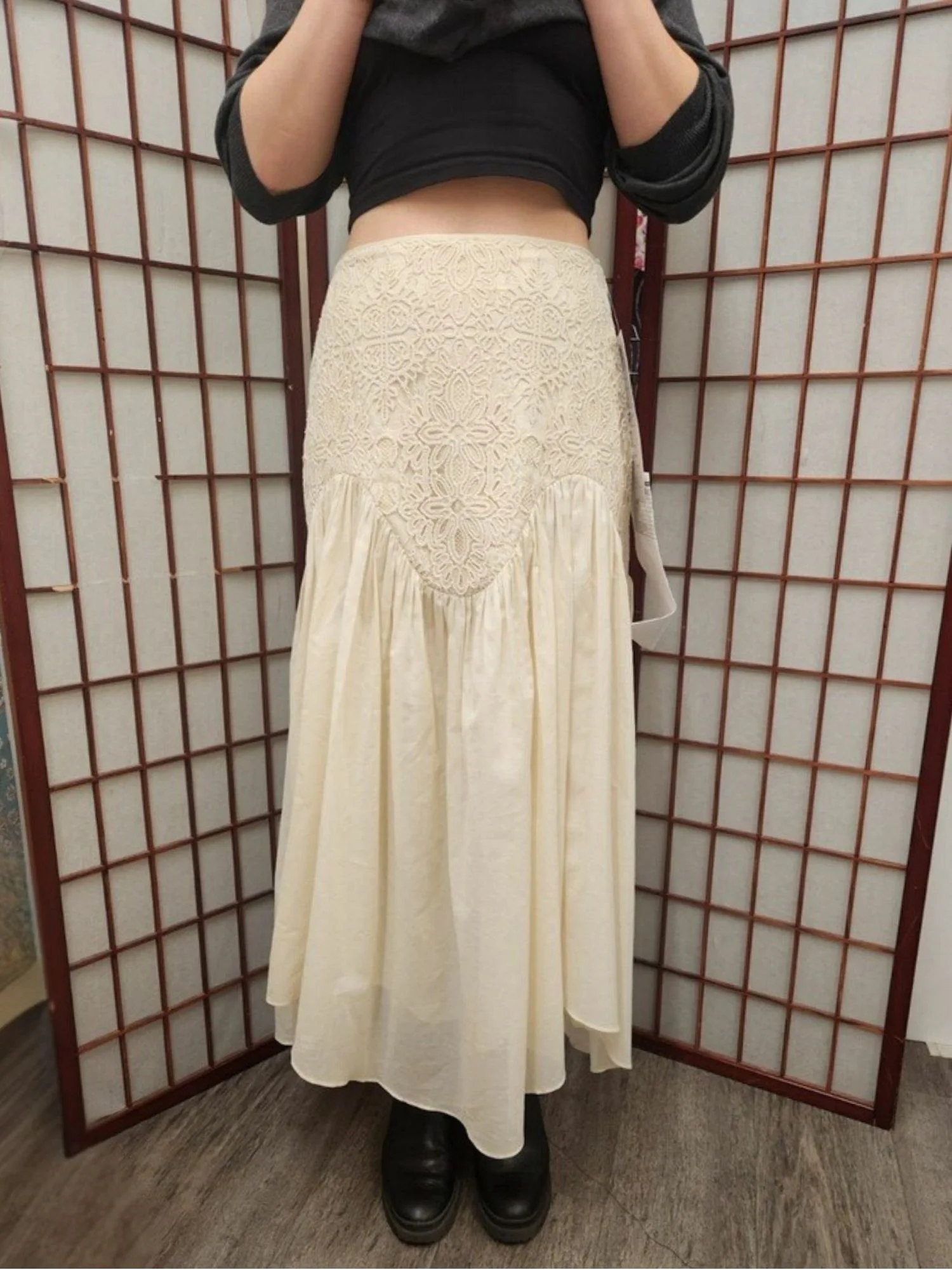 Person wearing a black top with three-quarter sleeves, a beige lace top with a floral pattern, and a long beige skirt with pleats, standing on a wooden floor in front of a folding wooden room divider.