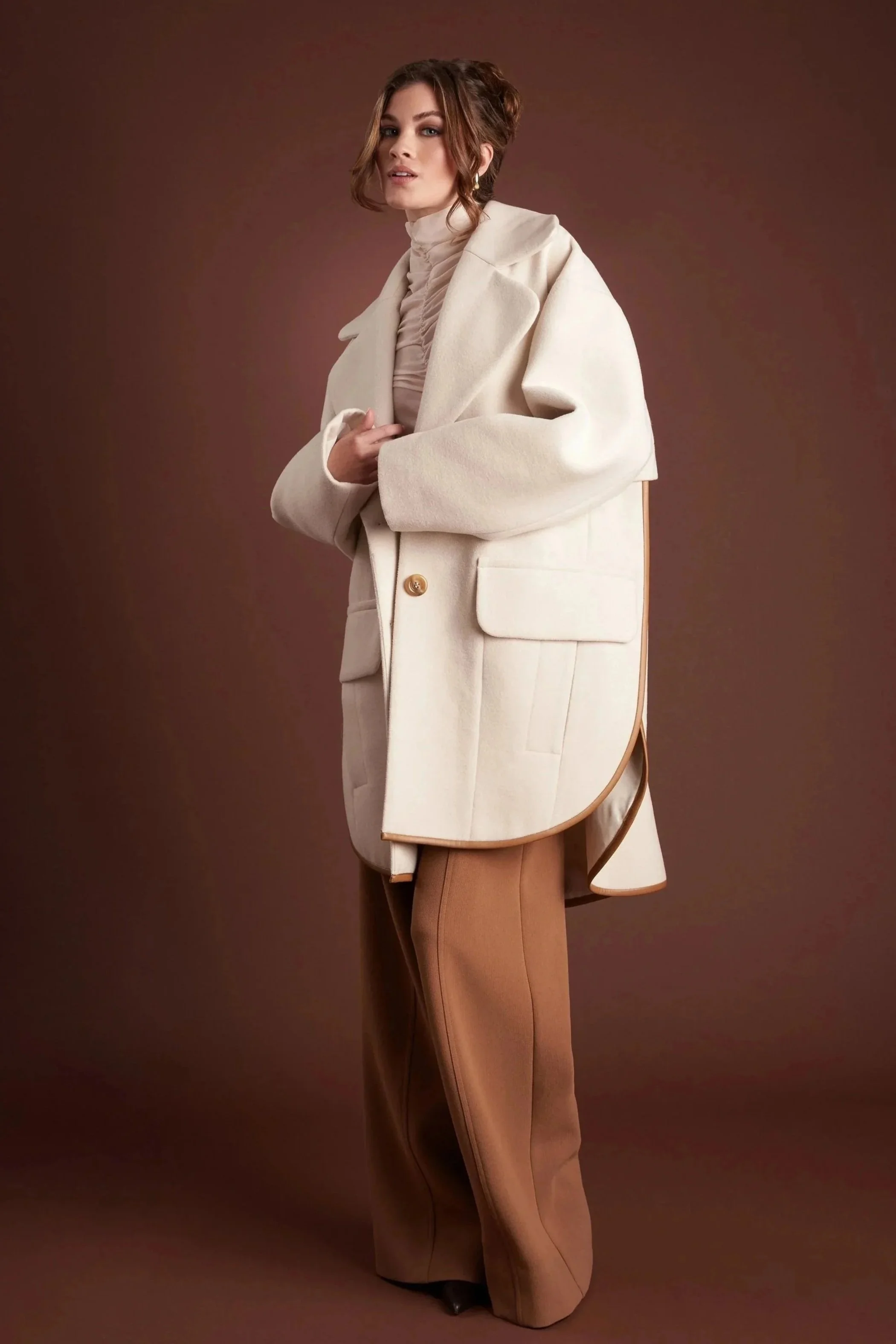 A woman in a cream-colored coat over a beige turtleneck, with tan wide-leg pants, posed against a brown background.
