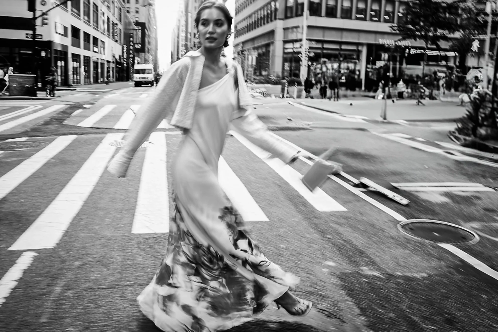 Black and white photo of a woman crossing a city street at a crosswalk, wearing a flowy skirt, a blouse, and sandals, with buildings and people in the background.