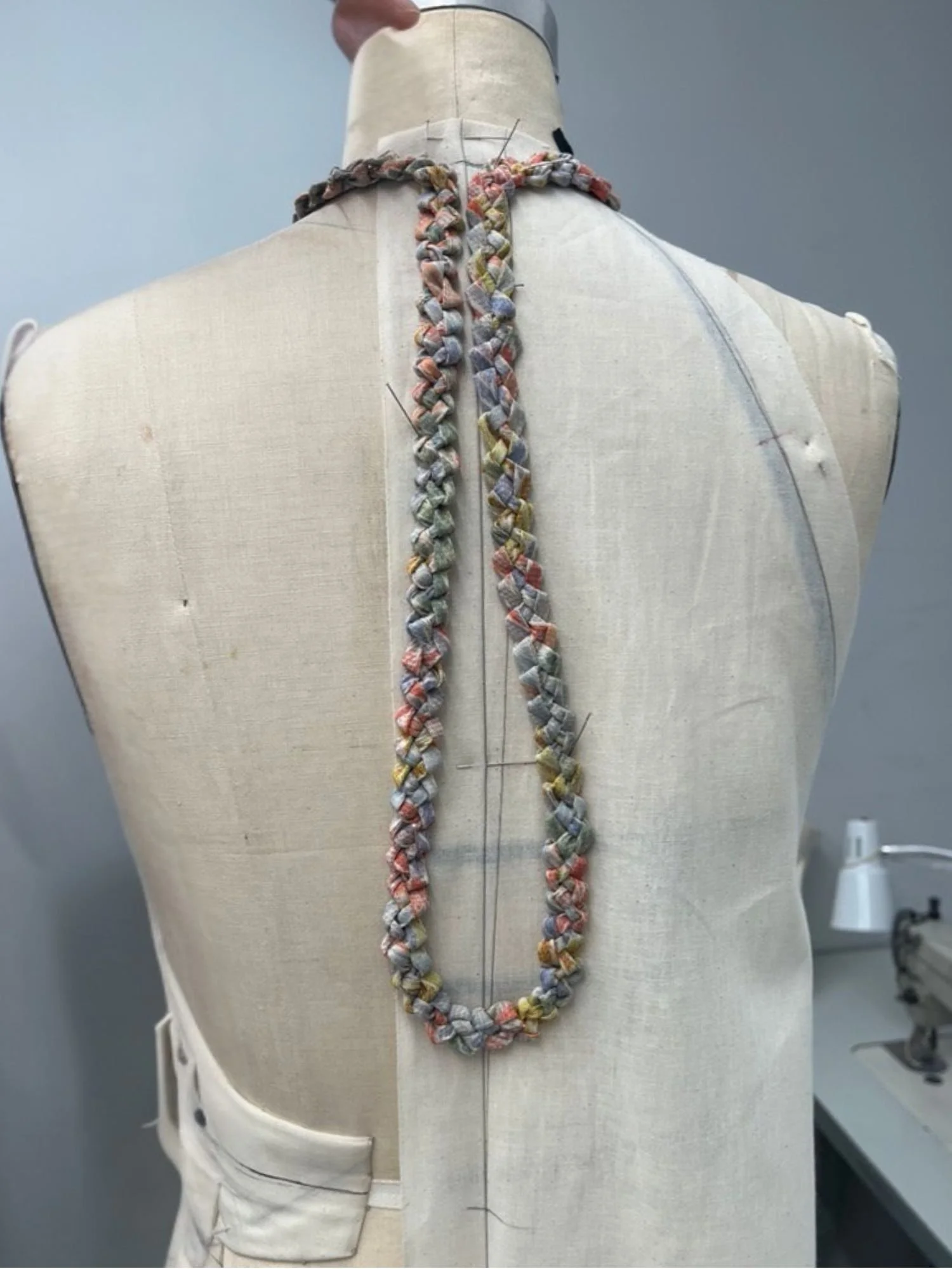A dress mannequin positioned in a tailoring or fashion design workspace wearing a colorful, braided, fabric necklace. The background includes a sewing machine and a lamp.
