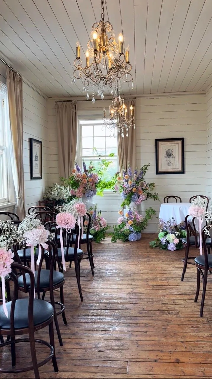Interior of a decorated room with chandeliers, flowers, chairs, and a table, likely set up for a wedding or special event.